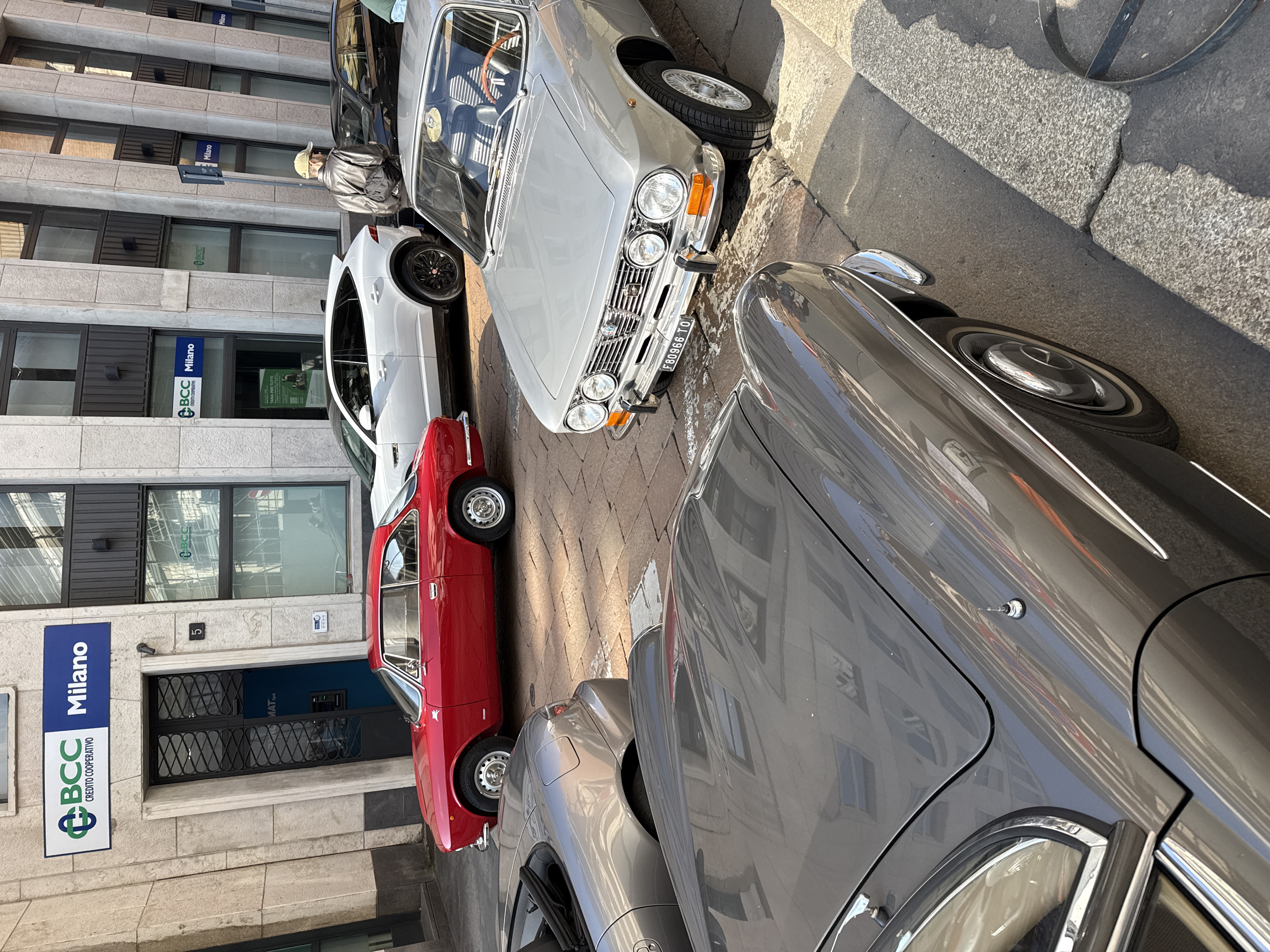 An urban street scene in Milan, Italy, depicts multiple parked vehicles on a stone-paved road under clear daylight.
The foreground is dominated by the hood and front fender of a dark grey classic convertible, possibly a Mercedes-Benz SL, reflecting the surrounding architecture. Behind it, a silver convertible, likely a Honda S2000, is partially visible.
To the right, a silver classic Alfa Romeo coupe is prominently parked, displaying an Italian license plate "F80966 TO" (Torino). Its interior features a wooden steering wheel and a yellow sticker on the dashboard; a hand is visible on the steering wheel. Behind this vehicle, a red classic Alfa Romeo coupe is parked, exhibiting a white 'biscione' emblem on its side. Further back, a white modern SUV is visible, followed by a partially obscured black modern vehicle.
The background features a multi-story building with light-colored stone cladding and dark-framed windows. Prominently displayed signs read "BCC Milano Credito Cooperativo." An entrance with a decorative metal grid door is marked with street number "5," revealing an ATM inside.
One male individual, wearing a light-colored baseball cap and a grey jacket with a black shoulder bag, stands facing away from the camera near the white SUV. The street surface is composed of irregular stone blocks, and the sidewalk is made of grey concrete or stone slabs. Strong sunlight creates distinct shadows across the scene.