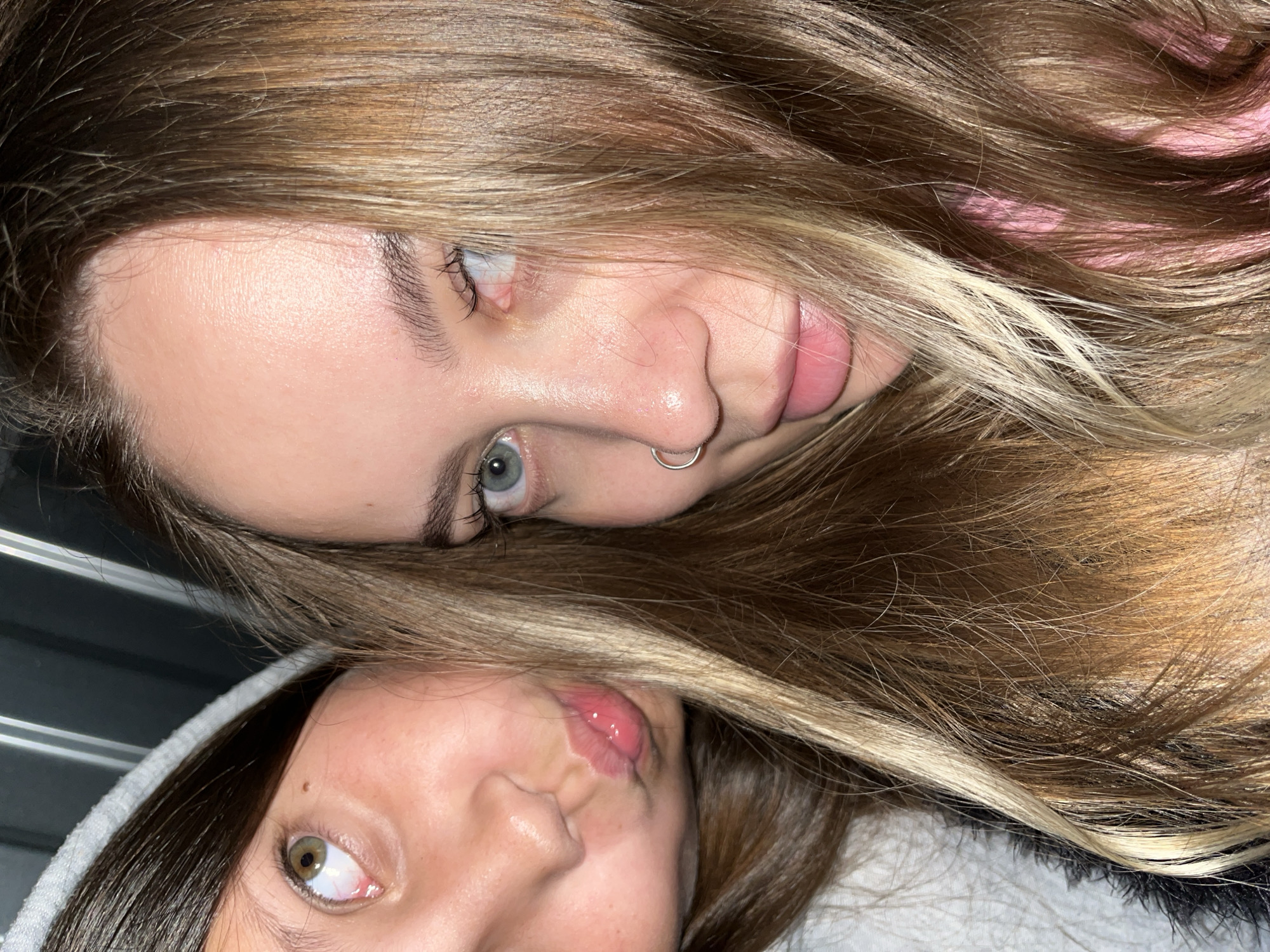 In this close-up selfie, two young females are positioned side-by-side, with their faces filling the frame. The female on the left, with brown hair and green eyes, is looking upwards and to the right. Her lips are slightly pursed, and she is wearing a grey hooded jacket. The female on the right has blonde and brown hair, with blue-green eyes and a silver hoop nose ring. She is looking directly into the camera. Her hair is partially obscuring her face. The background consists of dark, vertical lines. The image appears to have been taken indoors. There is no visible indication of the location being in Novopokrovskaya, Russia.