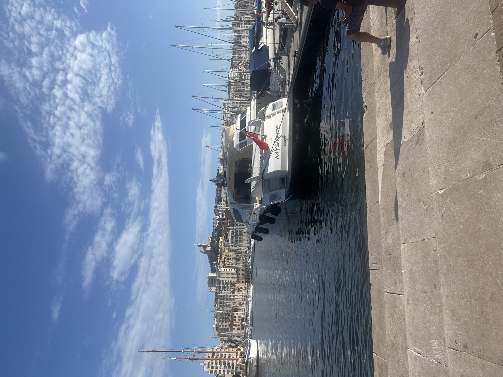 A sunny day shot captures a harbor scene in an unknown city. In the foreground, stone steps descend to the water's edge, where a person in shorts and a t-shirt sits on a bollard, casting a long shadow. The water is dark and slightly rippled. In the middle ground, a white motor yacht named "MYSTERE LONDON" is moored, its stern facing the viewer, with a red flag flying from its stern. Behind it, numerous other sailboats and motor yachts are docked, their masts creating a dense forest against the city skyline. The city itself is built on a hill, with numerous buildings and a prominent church or basilica at its peak. The sky is a bright blue, dotted with scattered white clouds.