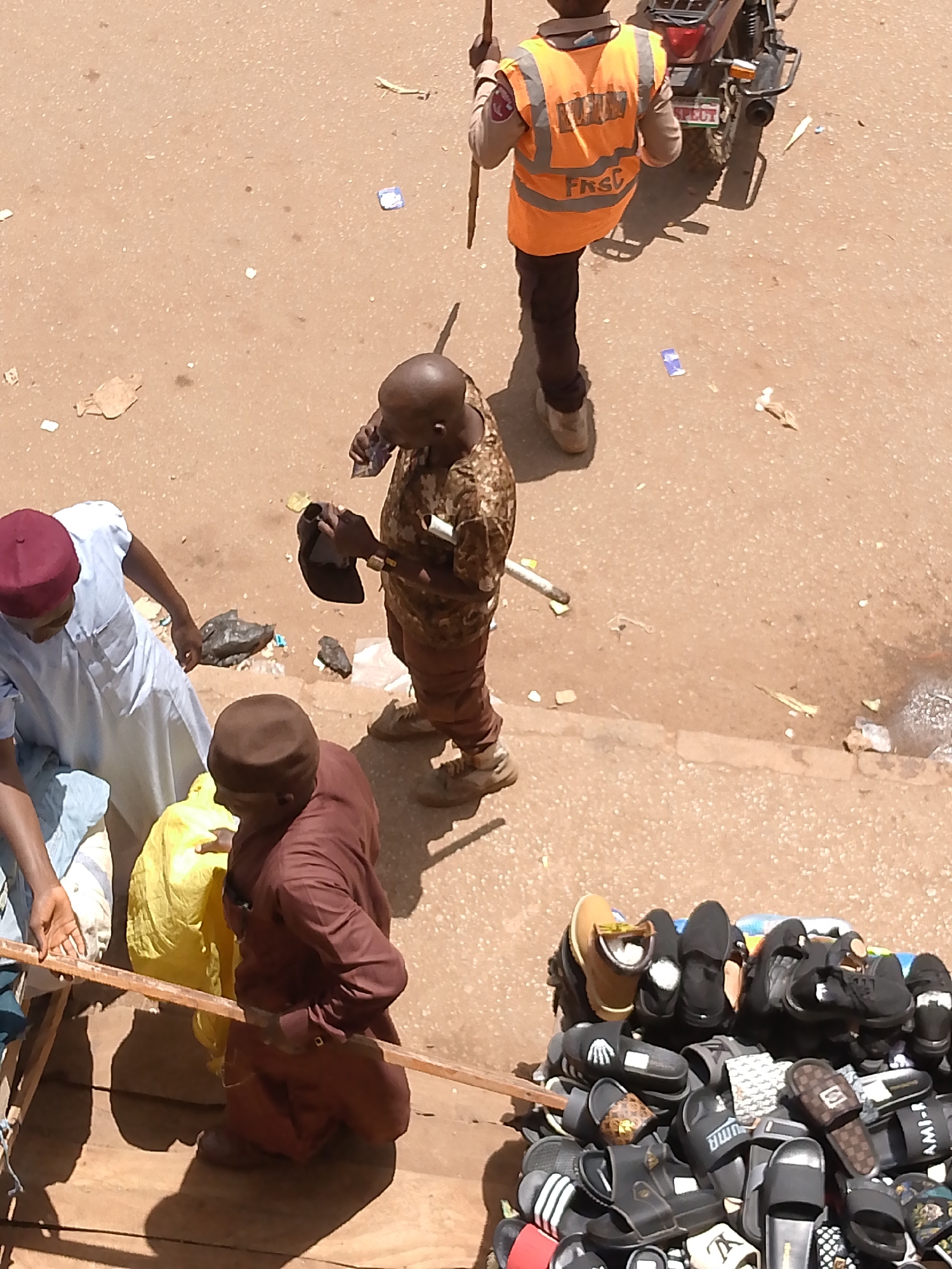 The image shows a street scene in Bukuru, Nigeria, from an elevated perspective. In the foreground, a stall displays a variety of sandals and slippers. A man in a brown robe stands nearby, with another man in a white robe and red hat partially visible to the left. Behind them, another man is wearing camouflage clothing and appears to be using a phone. Further back, a man in an orange vest labeled "FRSC" and dark pants stands near a motorcycle. Litter is scattered on the ground.