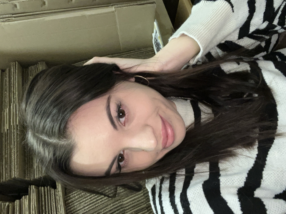 A young woman with long, dark hair is lying down. She is smiling slightly at the camera. She is wearing a black and white striped sweater. A gold hoop earring is visible on her left ear. The background is a stack of cardboard boxes. The lighting is soft and diffused.