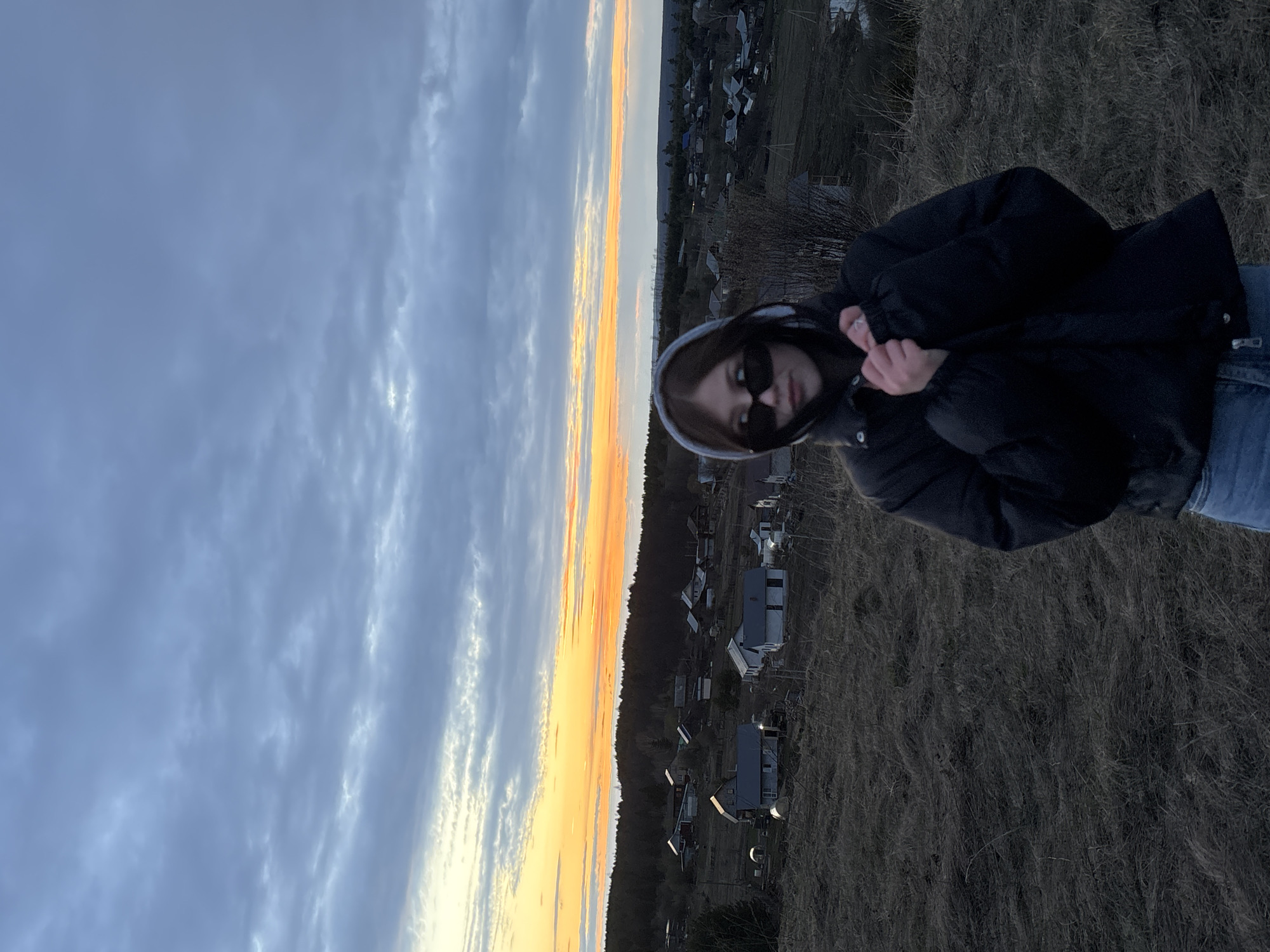 The image, taken in Фоки, Russia, captures a young person outdoors against a scenic backdrop, likely during either sunset or sunrise. The person is positioned on the right side of the frame, wearing a dark, puffy jacket with a light-colored hood pulled up, and dark sunglasses. Their hands are visible near their chest, appearing to adjust their jacket as they look towards the camera.
They are standing on ground covered with dry, brownish grass or sparse vegetation, suggesting a cooler season such as late autumn or early spring. In the background, a vibrant horizontal band of orange and yellow light from the setting or rising sun cuts across the horizon, separating the land from the sky. Below this illuminated horizon, a rural or semi-rural settlement with scattered houses and some trees is visible. The sky above the colorful band is mostly cloudy, displaying shades of blue and grey.
The primary activity appears to be posing for a photograph. There is no discernible text in the image. The overall impression is a serene outdoor moment, possibly capturing the transition of day to night or night to day.