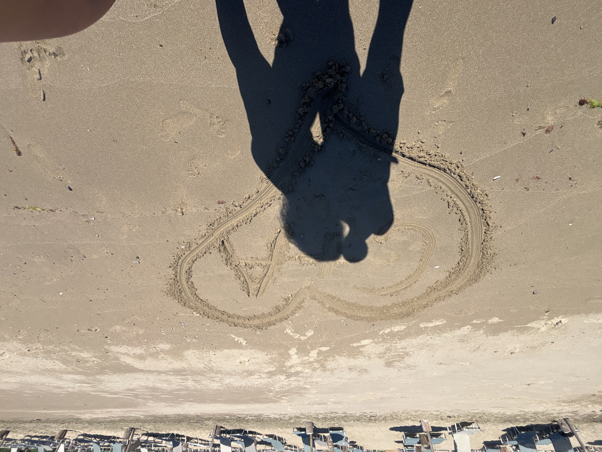 In Rezovo, Bulgaria, on a sunlit beach, a heart shape has been drawn in the sand. Inside the heart, the letters "C" and "A" are visible. The shadows of two people, one appearing to lean in for a kiss, are cast within the heart, obscuring the letters. In the background, rows of unoccupied beach chairs are arranged on the sand, with the sea not visible. The overall scene depicts a romantic moment captured on a beach.