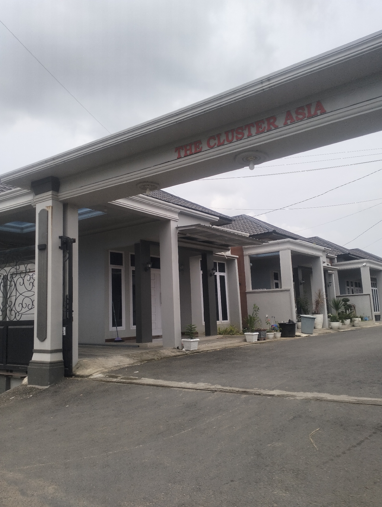 The image shows a wide shot of the entrance to a residential cluster named "THE CLUSTER ASIA". A large, grey, arched structure with decorative trim spans the road, bearing the name of the cluster in red lettering on its underside. The architecture of the houses visible beyond the entrance is modern and uniform, featuring grey walls and dark tiled roofs. Several houses are visible in a row, each with a covered porch supported by pillars. Potted plants and some outdoor furniture are scattered in front of the houses. The road in the foreground is asphalt, and the sky is overcast. There are no people or vehicles visible in the image.