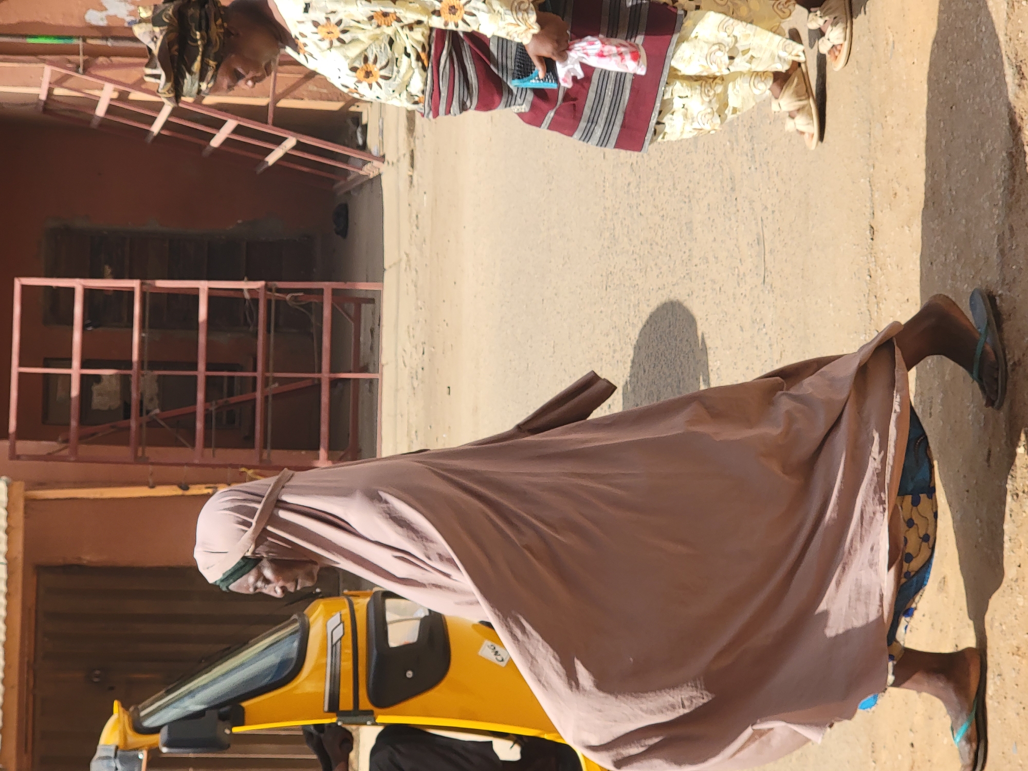 In Maiburiji, Nigeria, the image captures a scene with two distinct figures. One individual is adorned in a patterned yellow and white garment, accented by a red and gray striped shawl. They hold a blue object alongside a white and red patterned cloth. Another figure is enveloped in a brown fabric that obscures much of their form, with glimpses of blue patterned clothing visible beneath. This individual is walking near a yellow vehicle, partially visible in the frame. The background reveals buildings constructed with reddish-brown materials, featuring metal framework and windows. The ground is a light-colored dirt or concrete.