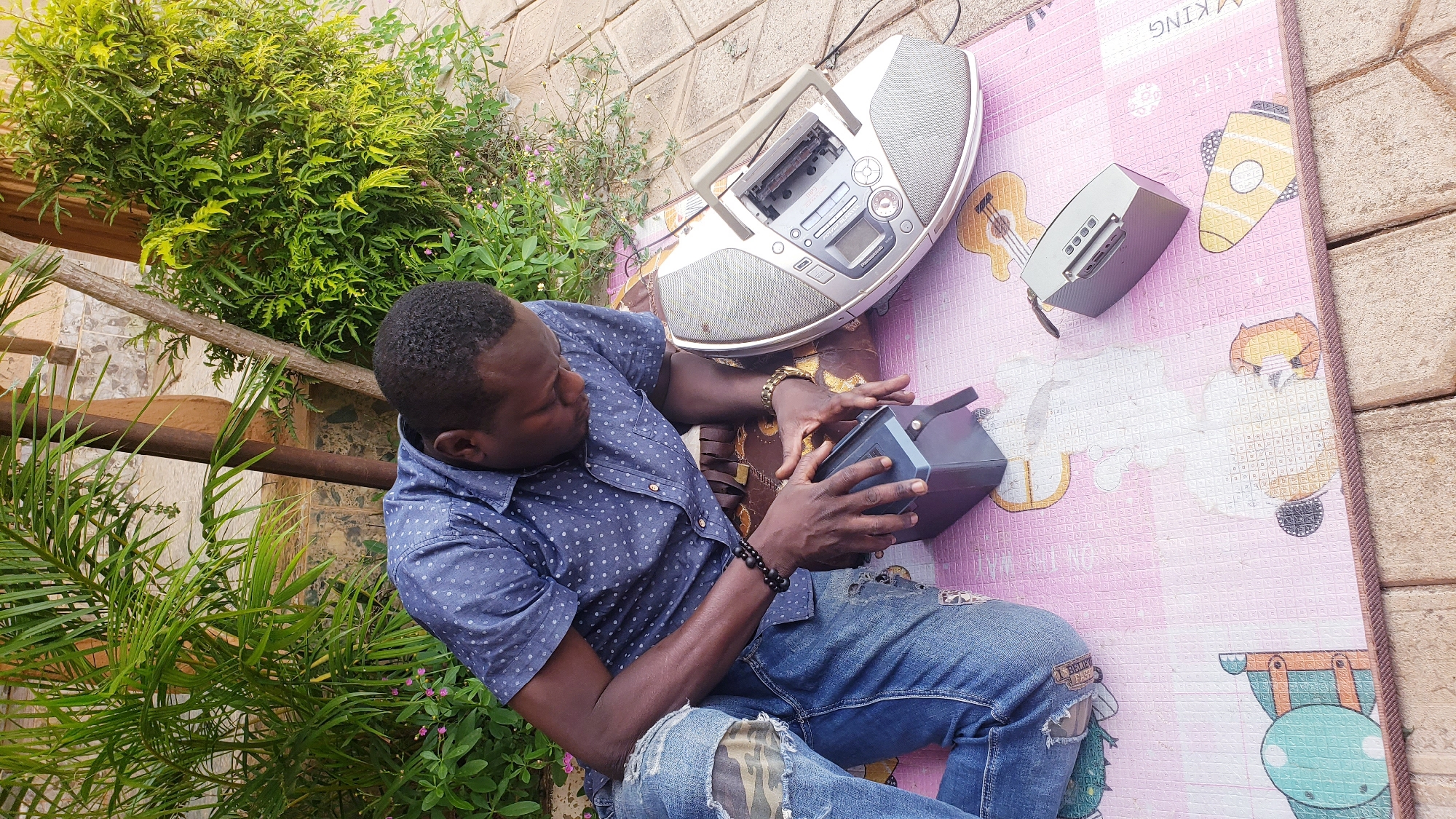 A Black man wearing a blue short-sleeved shirt with white polka dots and ripped blue jeans is kneeling on a pink mat on the ground in Sabon Tasha, Nigeria. He is looking down at a dark gray rectangular object, which he is holding with both hands. Next to him, to his right, is a silver boombox with a cassette deck. Further to his right is a smaller, silver portable speaker. The man has dark hair and is wearing bracelets on his left wrist. He is surrounded by lush green plants and foliage, with a cobblestone pathway visible in the background. The mat he is kneeling on has a colorful cartoon pattern.