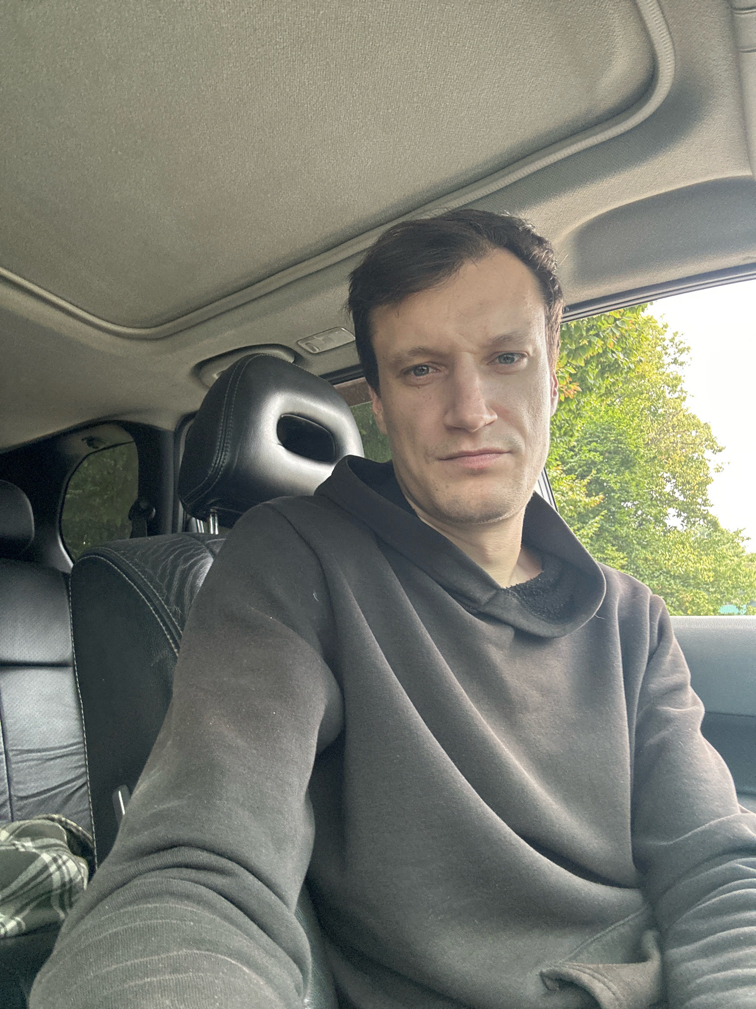 A man is sitting in the driver's seat of a car. He is wearing a dark gray hooded sweatshirt and dark pants. The car has black leather seats and a sunroof. Outside the car, trees are visible. The image was taken in Korchivka, Ukraine.