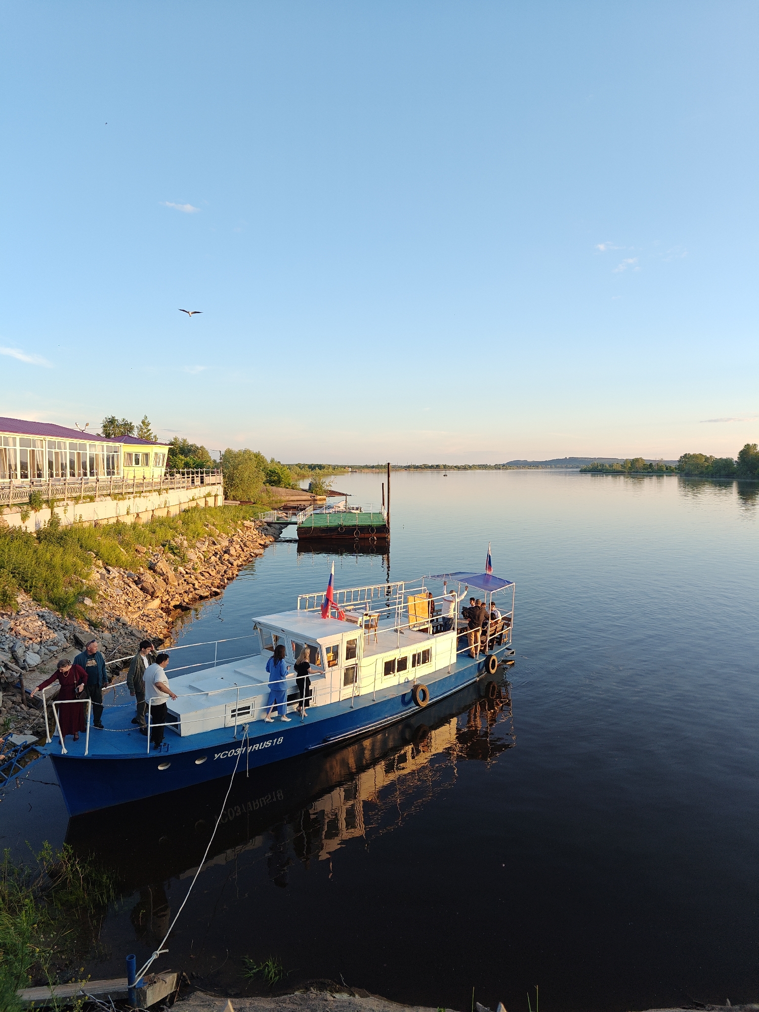This detailed description of the image is provided from a forensic, matter-of-fact perspective. The image depicts a wide river scene in Nikolo-Beryozovka, Russia, under a clear, light blue sky during what appears to be late afternoon or early evening. The lighting is warm and directional, suggesting golden hour, with the sun illuminating the left riverbank and parts of the boat. In the upper portion of the frame, the sky is a gradient of light blue, slightly lighter towards the horizon. A single, small, distinct white cloud is present in the upper-center, with a few faint wisps of clouds visible elsewhere. A dark bird with spread wings is captured in flight in the upper-left quadrant, with a smaller, barely visible speck in the sky further left. The majority of the lower half of the image is occupied by a wide, calm river with dark blue-green water, reflecting the sky and the warm sunlight, particularly near the foreground. Prominently featured in the mid-ground is a leisure boat with a blue hull and a white cabin, docked close to the left bank. A white rope tethers the boat to a small post on the bank. The registration "УС0231RUS18" is clearly visible in white lettering on the blue hull near the bow. Several black tires serve as fenders along the boat's side. The boat's reflection, including the hull, cabin, and the inverted registration, is distinctly visible in the still water. A Russian flag (white, blue, red horizontal stripes) flies from a pole at the stern of the boat. The upper deck features a blue canopy structure. Approximately eight to ten individuals are visible on and around the main boat. Three people, two men and one woman in a maroon dress or coat, are standing on the boat's main deck near the bow, facing the bank, with the woman holding onto a railing. On the upper deck under the blue canopy, several other people are present, including two women in blue and black attire standing on the starboard side. On the rocky bank directly adjacent to the boat, a man in a dark jacket stands, appearing to interact with those on board. The left riverbank, sloping towards the water, is covered with green vegetation, tall grasses, and exposed rocks, all strongly lit by the warm, golden sunlight. Further down this bank, a light yellow building with numerous large glass windows and a distinctive purple roof