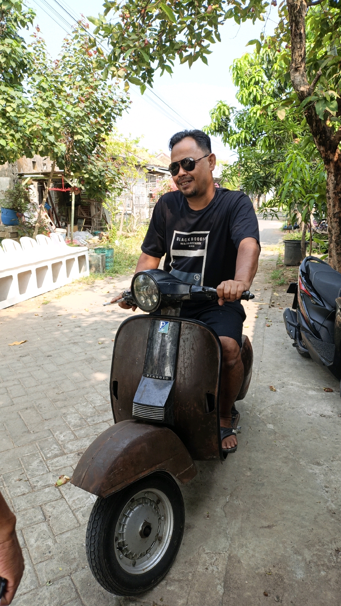A man wearing sunglasses, a black t-shirt with a white graphic, and black shorts sits on a rusty brown Vespa scooter. He is outdoors, with lush green trees and a bright blue sky in the background. The scooter is parked on a paved pathway. To the right of the scooter, another black motorcycle is partially visible. In the background, there are buildings, a fence, and potted plants. The overall scene suggests a warm, sunny day in what appears to be a residential area in Cikal, Indonesia.