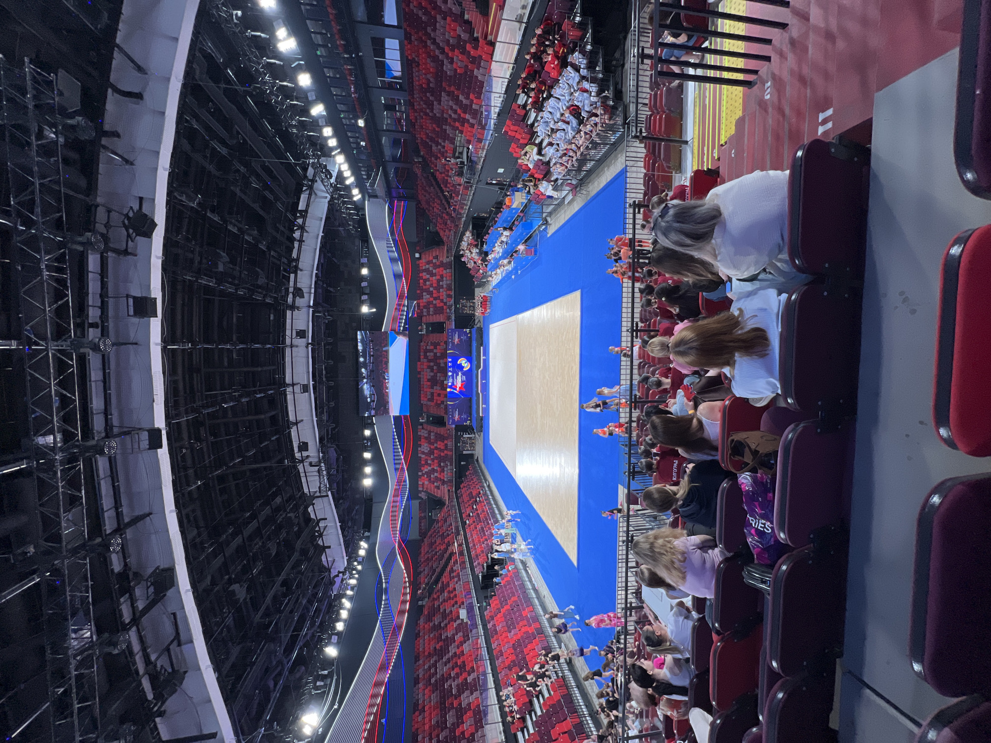 The image shows an indoor arena with tiered seating. The arena floor is a blue mat with a large beige rectangular area in the center, likely a competition surface. Several young people, possibly athletes, are standing and walking on the blue mat. Rows of empty red seats are visible in the foreground and on the sides of the arena. Some spectators are seated in the lower rows of the arena, facing the competition area. The arena has a complex ceiling structure with lighting and support beams. The overall impression is that of a sporting event or performance taking place in a large, modern venue.