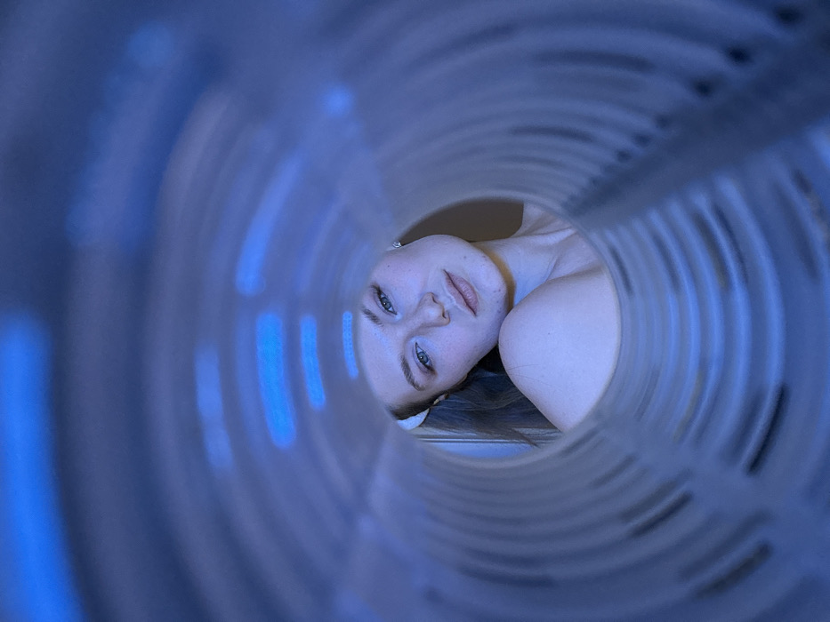 A woman's face is framed within a cylindrical, tunnel-like structure. Her gaze is directed toward the viewer, creating an intimate and slightly surreal perspective. The structure surrounding her face features repeating circular patterns, creating a sense of depth. The lighting is predominantly blue, lending the image a cool and ethereal quality. Her skin is fair, and her eyes are striking, drawing the viewer's attention to her expression.