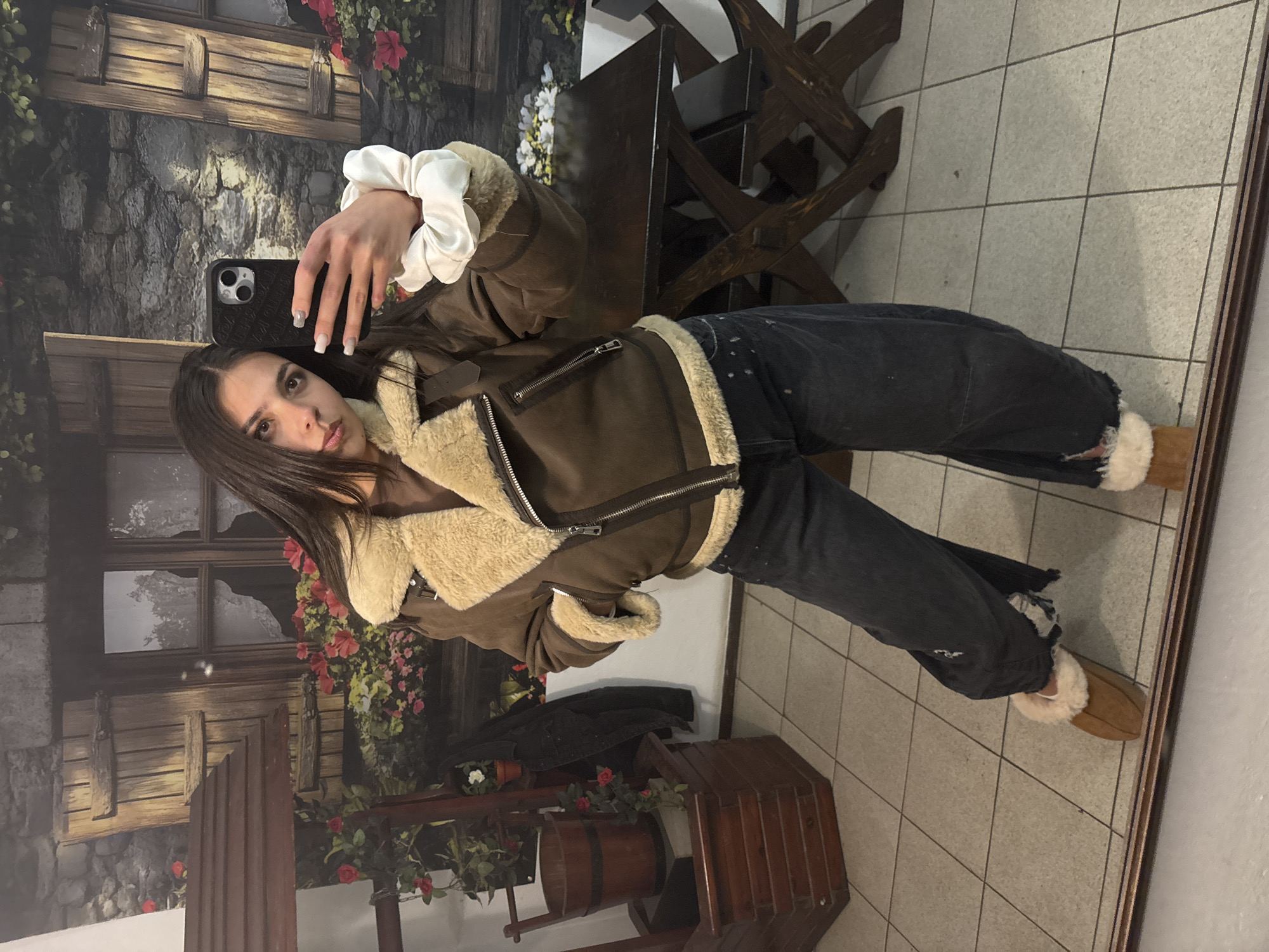 A young woman is taking a selfie in a room with a unique backdrop. She holds a smartphone in her right hand, a white scrunchie around her wrist. She wears a brown aviator-style jacket with a cream-colored shearling lining and dark-wash denim jeans, along with brown slippers with shearling trim. The background features a painted mural or wallpaper depicting a rustic village scene with a stone building, flower boxes, and wooden elements, all contributing to an idyllic, possibly European countryside motif. Additional furniture includes a dark wooden table and chairs. The flooring is tiled in a light color. The image captures an informal, self-portrait moment within an enclosed setting. The location is not Gabrovo, Bulgaria.