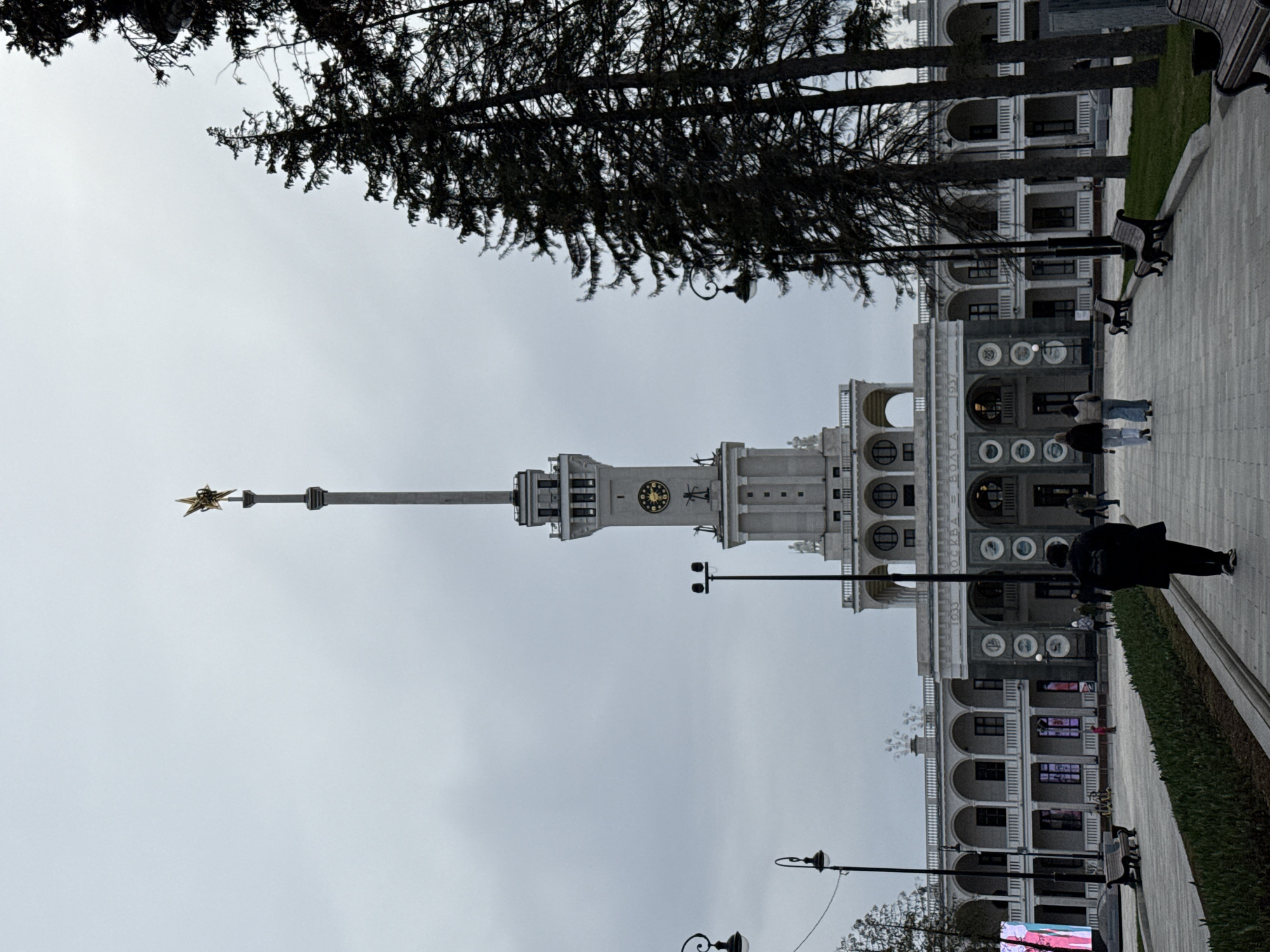 The image displays a grand architectural complex, likely a river terminal or a public building, captured under an overcast sky. The photograph is oriented so that vertical elements appear angled.
The central subject is a multi-story building constructed in a classical or Stalinist Empire style, characterized by numerous arched windows, columns, and ornate architectural details. A prominent clock tower rises from the building's center, featuring a dark clock face with golden numerals. A tall spire extends from the tower, topped with a distinct golden five-pointed star.
In the foreground, a wide paved walkway runs alongside the building, bordered by a strip of green grass and several benches. Mature trees with dark, dense foliage are visible at the top of the frame, with their trunks appearing on the left side of the image. Several lampposts are spaced along the walkway.
A few individuals are present, providing a sense of scale. One person, dressed in a dark coat and carrying a backpack, walks along the paved area. Two other people stand closer to the building in the mid-ground.
Notable details include the overcast sky, indicating a cloudy daytime setting. Visible text on the building's facade, located below the clock tower, reads "МОСКВА" (Moscow), "ВОЛГА" (Volga), and the year "1937." These inscriptions, along with the distinctive architecture and the golden star, strongly suggest the location is the Moscow Northern River Terminal (Severny Rechnoy Vokzal). The scene appears calm, depicting typical public activity in a large, historic space.