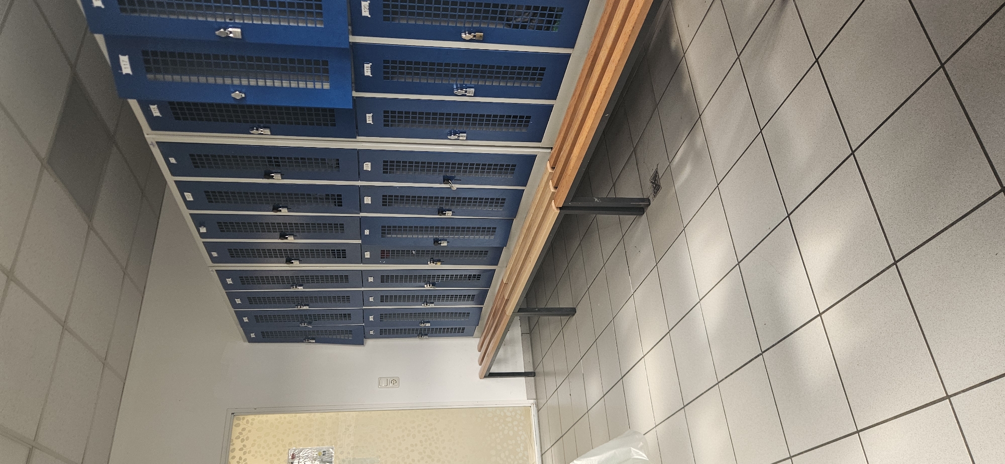 The image depicts an interior scene, likely a locker room. A wall of blue lockers with metal grills is visible, running along the length of the room. Adjacent to the lockers is a wooden bench with metal supports. The lockers appear to be secured with various types of locks. The floor is covered in square tiles with dark grout lines. The lighting is diffused, and no people are visible. There's a door or window visible at the end of the locker row. The image was taken in Delbrück, Germany.