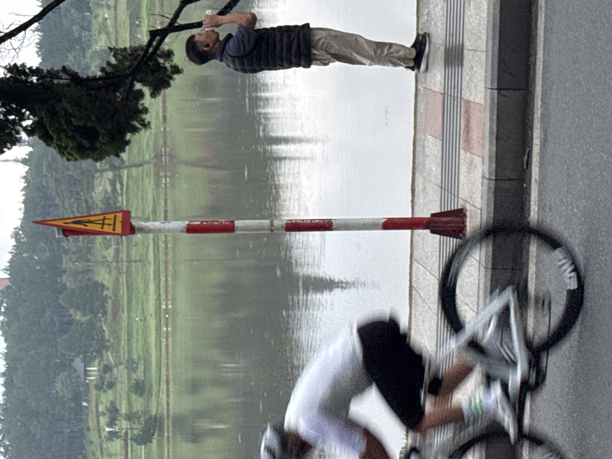 The image captures a scene by a lake in Da Lat, Vietnam, likely Xuan Huong Lake, a popular tourist destination known for its walking paths and recreational activities. In the foreground, a cyclist wearing a white t-shirt, black shorts, and a helmet is blurred due to motion, indicating they are actively riding. To the right, a man stands on a paved walkway, wearing a dark vest over a polo shirt and light-colored pants. He is holding a white cup to his mouth, appearing to drink. Behind him, a red and white striped pole with a yellow triangular warning sign featuring a pedestrian symbol stands near the walkway. The lake occupies the mid-ground, reflecting the overcast sky and the treeline along its edge. In the background, a park area with scattered trees and a small gazebo is visible. The overall atmosphere suggests a cool, possibly damp day given the overcast conditions and the lush greenery.