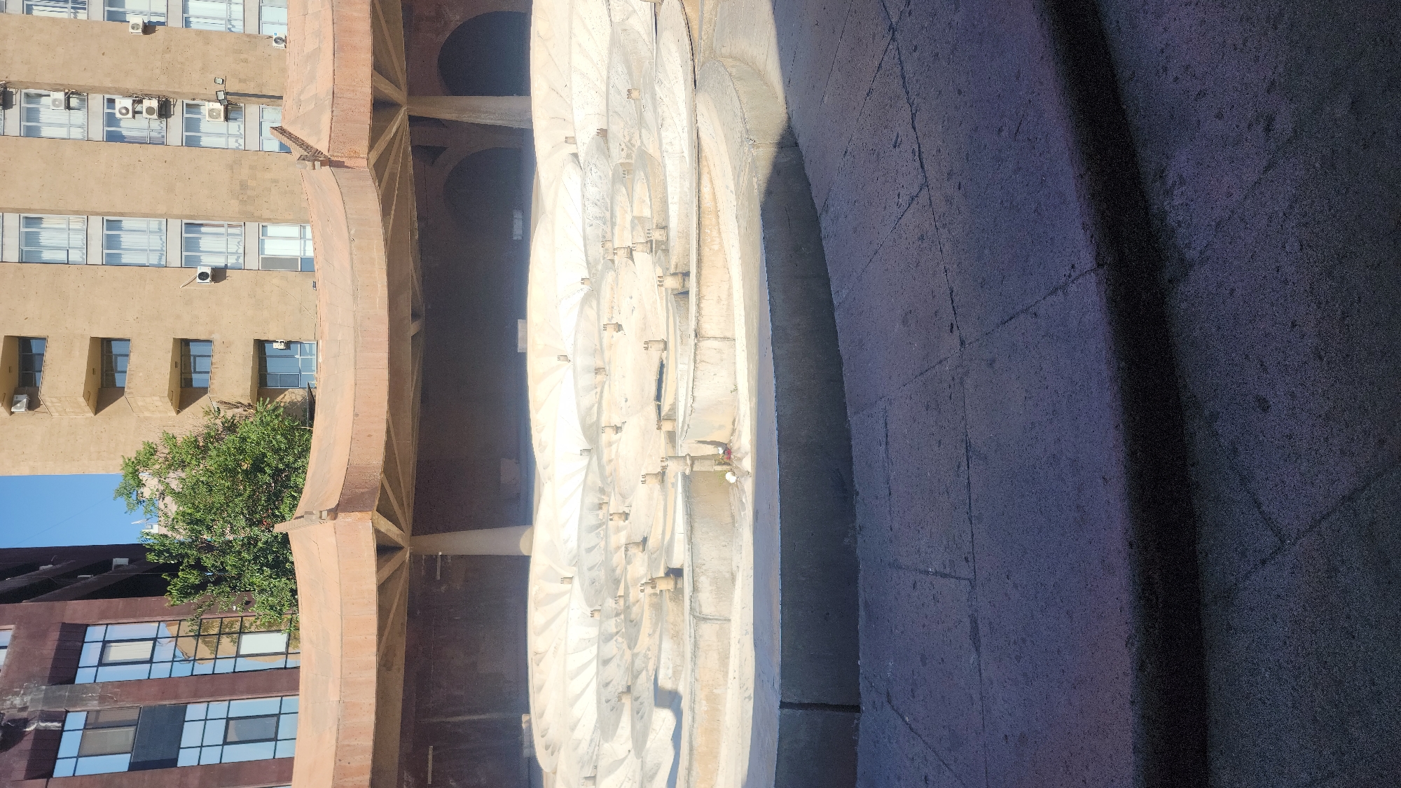 The image shows a view of the Cascade complex in Yerevan, Armenia, looking up towards the top of the monument. In the foreground, there is a paved walkway with dark granite slabs. Behind this, a large, multi-tiered dry fountain is visible, constructed from concrete and featuring numerous small spouts. The fountain area is surrounded by low concrete walls and curves. Above the fountain, a wide, orange-colored canopy supported by pillars extends across the scene. In the background, two buildings are visible: a darker building with large, reflective windows on the left, and a lighter, beige-colored building with a grid of windows and several air conditioning units on the right. Green trees are interspersed between the buildings. The sky is a clear, bright blue. There are no people or animals in the image.