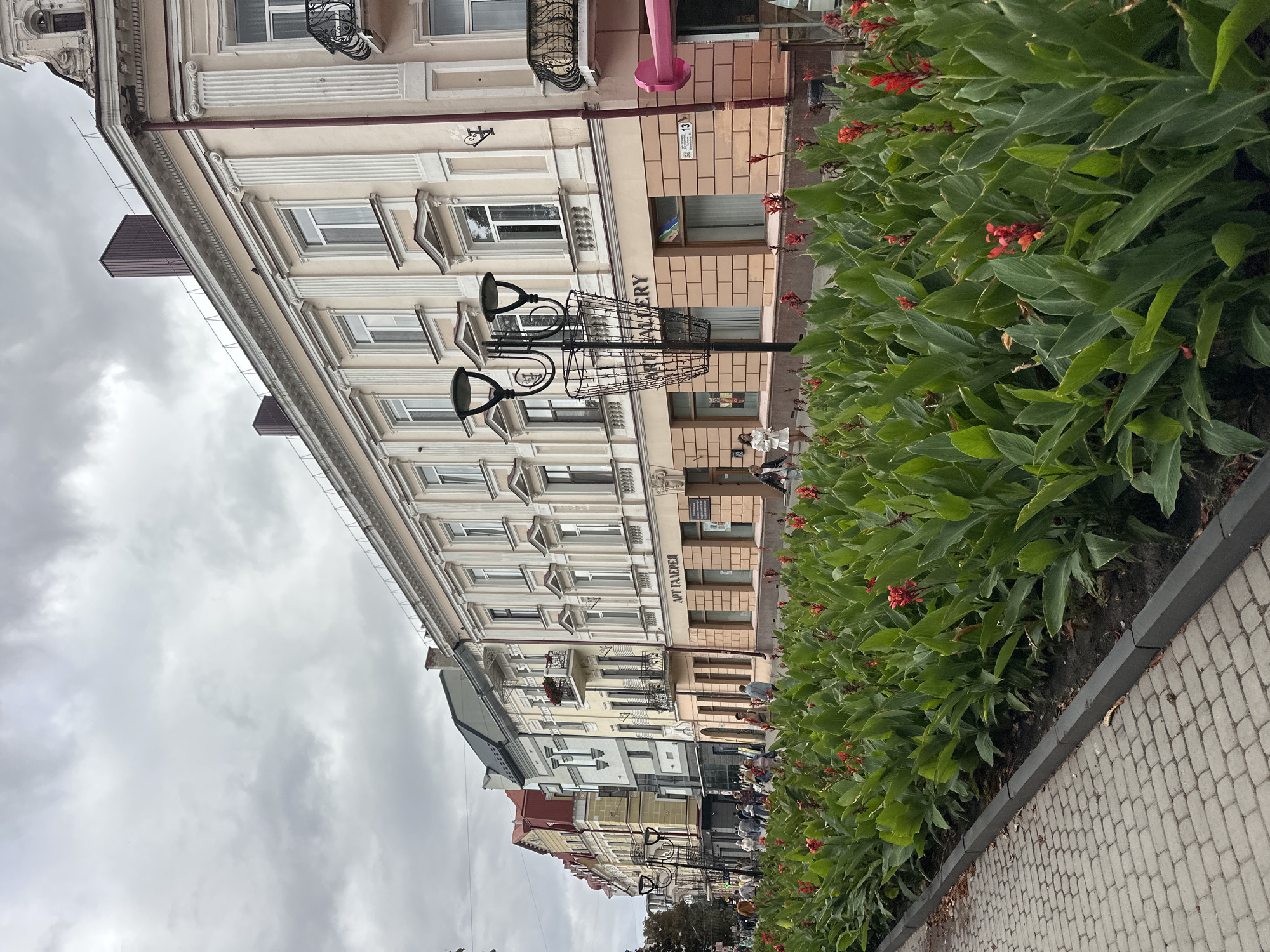The image depicts a street scene in Ternopil, Ukraine, with a prominent row of tall green plants with red flowers lining a cobblestone sidewalk. Behind the plants, a row of multi-story buildings with ornate architecture stands. The buildings are painted in light colors, with decorative elements like columns and balconies. Signs on the buildings indicate businesses, including one that reads "ART GALLERY" in Cyrillic script. Several people are walking on the sidewalk and in front of the buildings. The sky is overcast, suggesting a cloudy day. The scene conveys a sense of urban life and architectural heritage.