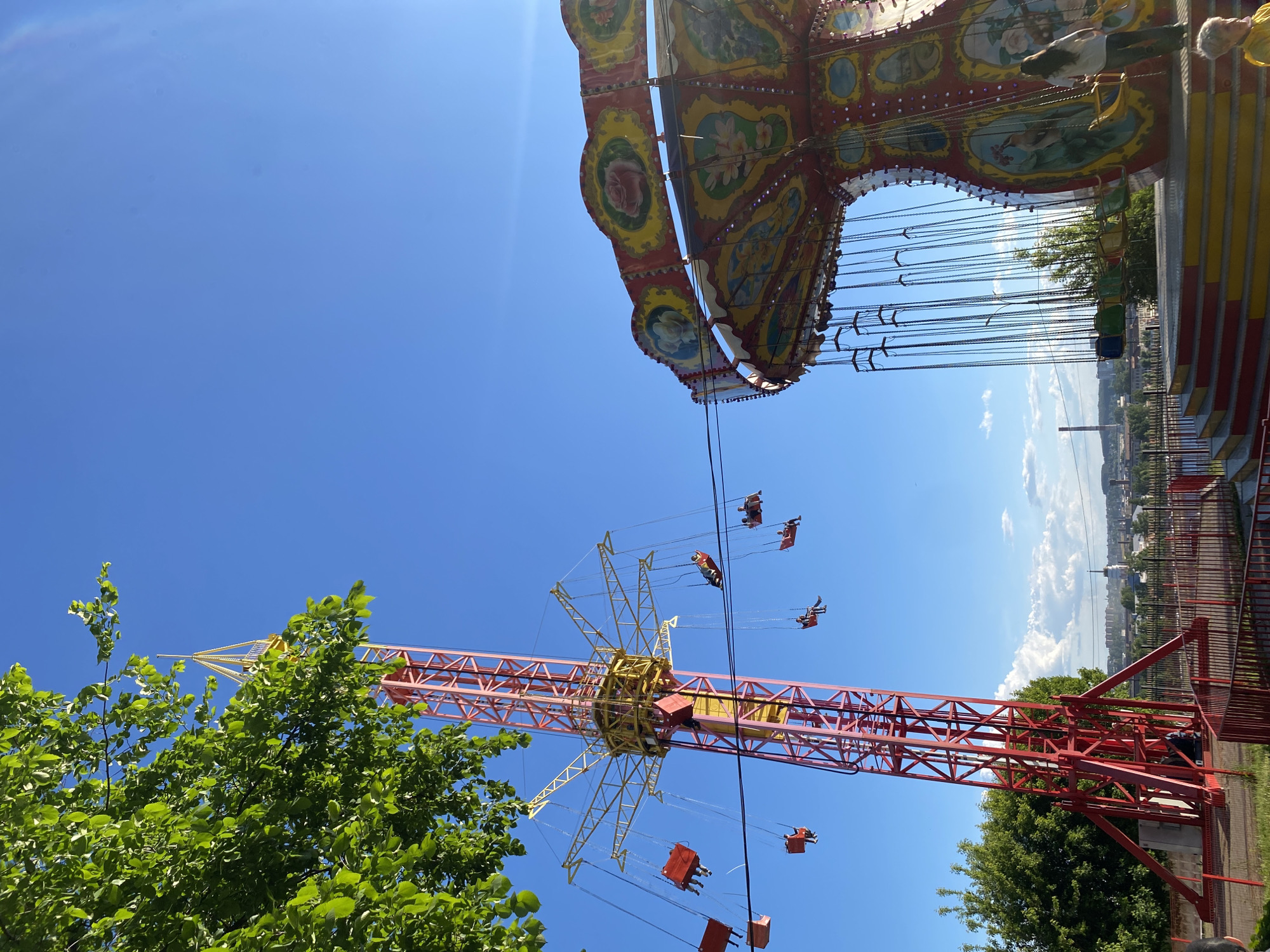 This image, captured in Izhevsk, Russia, depicts a bustling amusement park scene on a bright, clear day. The photograph is taken from a low angle, looking upwards towards the sky and the towering structures of two distinct swing rides.
In the upper left quadrant, lush green tree foliage provides a natural foreground element. Extending from the upper left towards the bottom right of the frame is a tall, modern swing ride featuring a robust red and yellow metal framework. Its central mechanism, also in yellow, shows radial arms from which numerous individual swing seats are suspended by chains. Several people are visible in these seats, soaring through the air, indicative of the ride being in full operation.
Dominating the mid-right side of the image is a second, more traditional and ornate swing ride canopy. This large, circular structure is richly decorated with intricate patterns in red, yellow, and green, adorned with floral motifs such as roses and other blossoms. Many chains hang from its underside, leading to multiple swing seats, some of which are visible with riders. One person is also partially visible on the platform above the canopy.
The sky is a brilliant, deep blue with only a few scattered wispy clouds, suggesting excellent, sunny weather. In the background, particularly visible beneath the ornate canopy and extending to the horizon, is a distant cityscape or townscape. This panoramic view implies that the rides offer expansive vistas of the surrounding area. The strong sunlight indicates it is likely midday or early afternoon. No discernible text is visible in the image. The overall atmosphere conveyed is one of joy and excitement at a lively fairground.