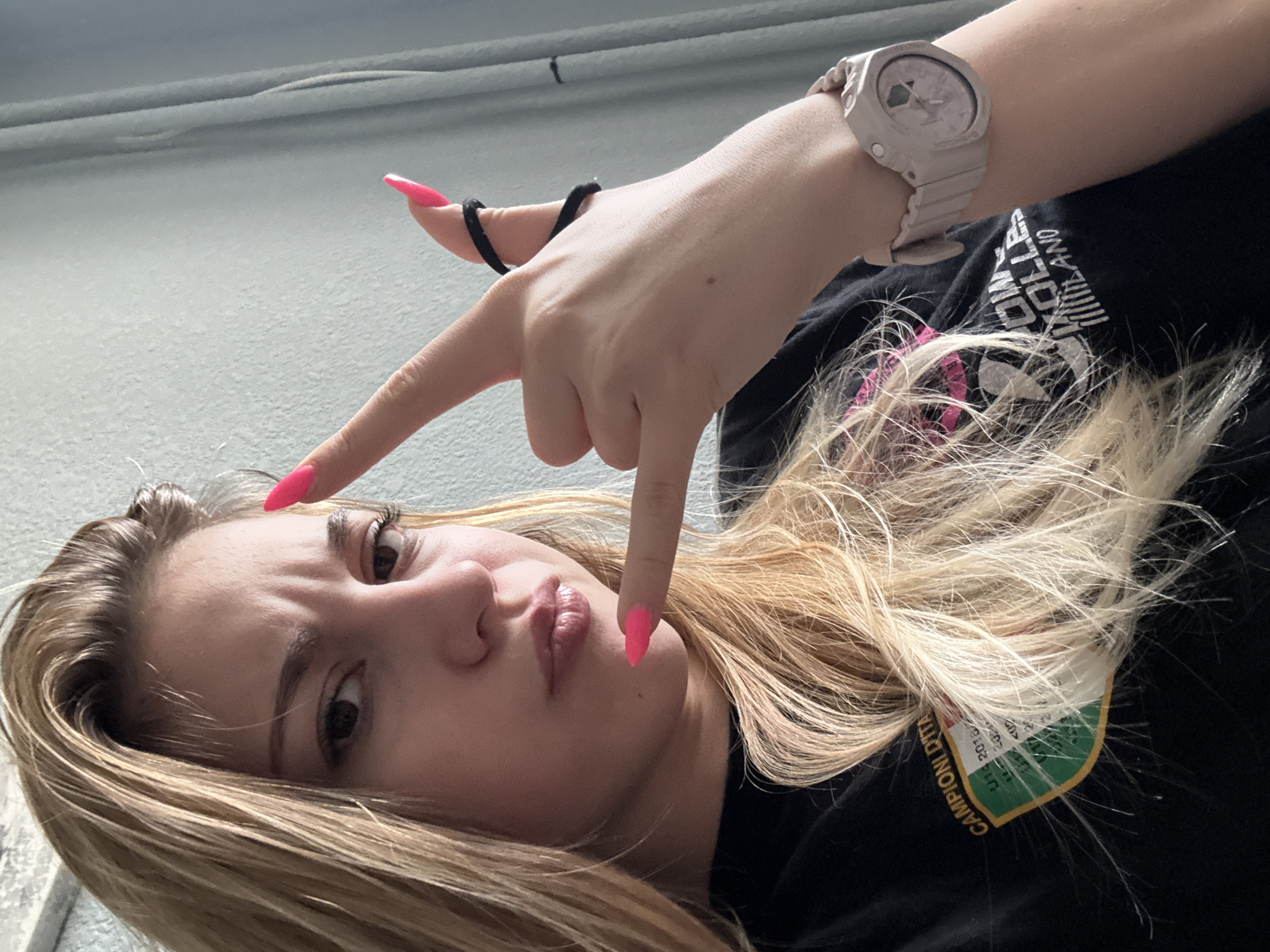 A young woman with blonde hair and pink nail polish poses for a selfie in what appears to be an indoor setting with light gray walls. She is making a hand gesture with her fingers pointing downwards and outwards. A watch is visible on her wrist. She is wearing a black t-shirt with white lettering that partially reads "ROLLER". A patch with the words "CAMPION DITA" is visible on the shirt. The location in Sofia, Bulgaria, is not discernible from the image.