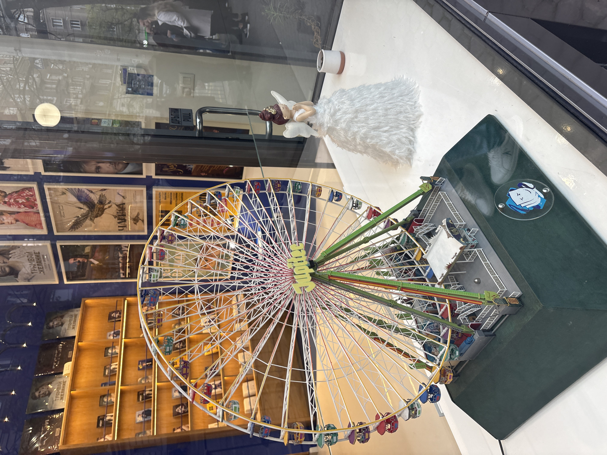 This media file captures a detailed display within what appears to be a shop or cafe window, seen from a slightly elevated and angled perspective. The primary subjects are a miniature Ferris wheel model and an angel figurine.
The Ferris wheel, centrally located, features a yellow hub with "JUMBO" visible, white support beams, green connecting rods, and numerous multi-colored passenger cabins. It rests on a dark green rectangular base. Behind the Ferris wheel, an angel figurine with a white, fluffy dress, brown hair, and wings is positioned on the white display surface, alongside a small white cup or pot.
The glass surface of the window presents multiple reflections. These include glimpses of an urban exterior with buildings, trees, and a person in light clothing. Visible in reflection, on a door handle, is the word "PULL". The interior space behind the display shows a wall adorned with framed posters or artwork. Recognizable text on these posters includes "ЧАЙКА" (Chayka, meaning Seagull) and multiple instances of "ТРАМВАЙ ЖЕЛАНИЕ" (Tramvai Zhelanie, meaning A Streetcar Named Desire). Below these posters, wooden shelves filled with various items, possibly books or other merchandise, are visible. A small, clear, circular coaster-like object with a blue logo, depicting a stylized figure, rests on the green base of the Ferris wheel.
The scene is brightly lit by natural light, indicating it is daytime. No active event is taking place; it is a static display.