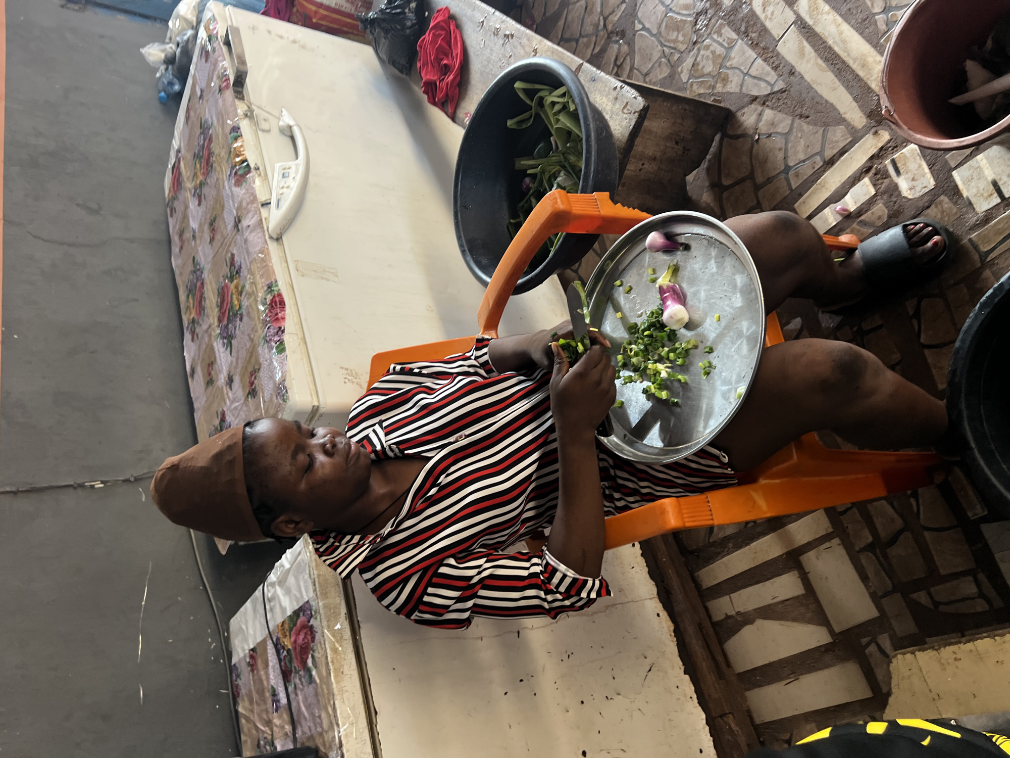A medium shot captures a woman in Nkpor, Nigeria, seated on an orange plastic chair. She wears a brown head covering and a striped shirt with red, white, and black vertical lines. The woman is actively chopping green vegetables and red onions on a circular metal tray held in her lap. Beside her, a large black plastic bowl contains more green vegetables. In the background, a white chest freezer is draped with a floral patterned cloth. A wooden surface holds a red cloth and a black plastic bag. The floor is tiled with a pattern of light and dark rectangular tiles, and a brown bucket is visible in the foreground.