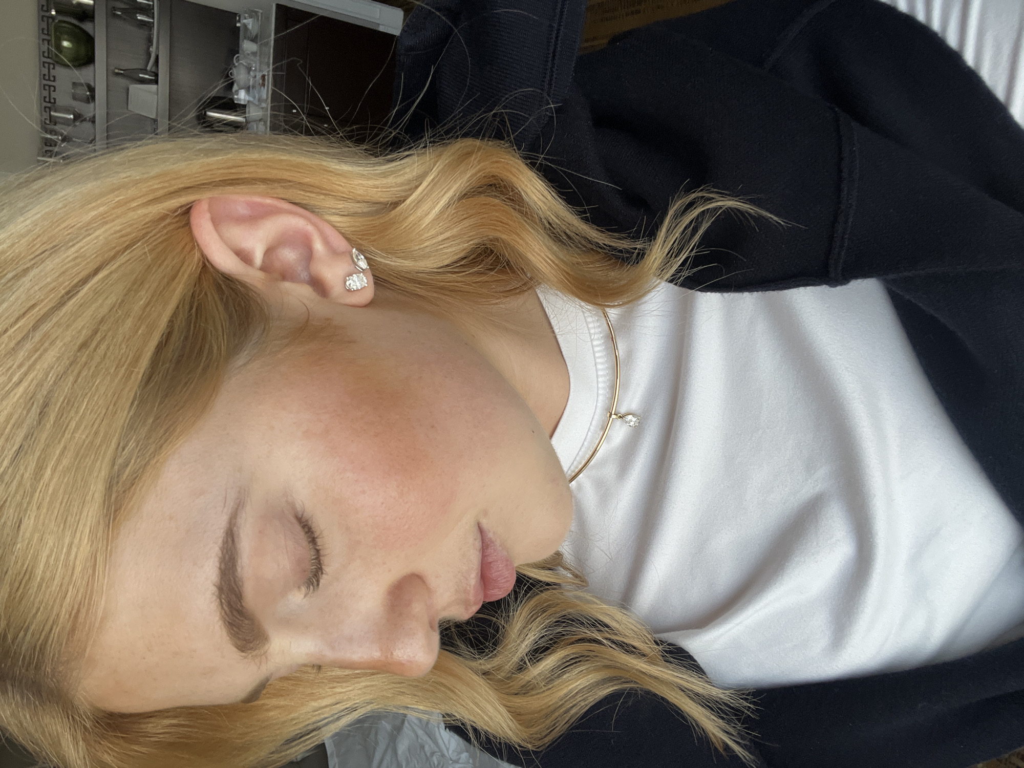 A close-up shot shows a person with blonde hair, wearing a black hooded sweatshirt over a white t-shirt, and gold jewelry. The person's eyes are closed and their head is tilted to the side. The background is indistinct, suggesting an indoor location.
