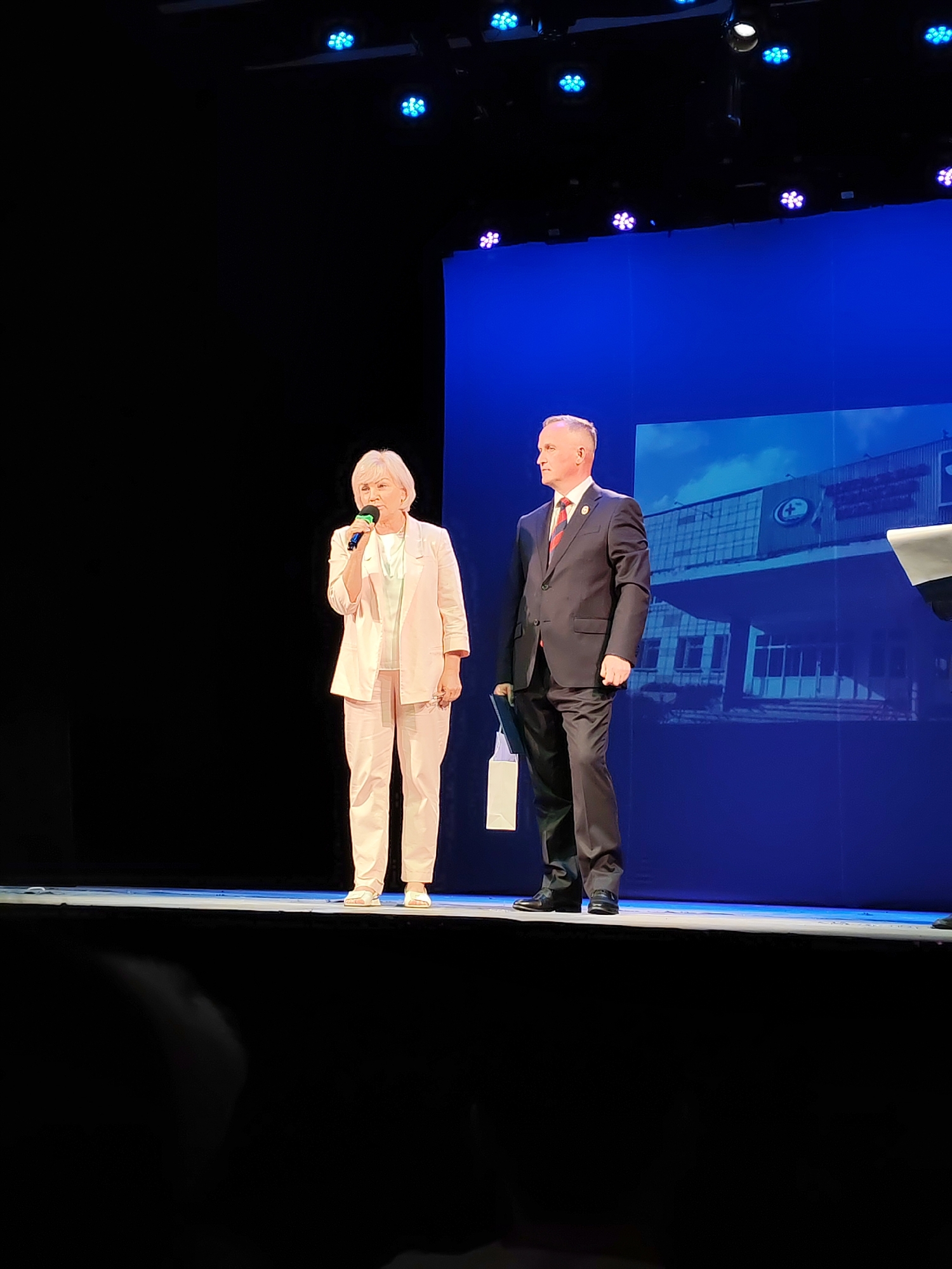 Two people, a woman and a man, stand on a stage in front of a large blue screen that displays a building. The woman is on the left, wearing a light-colored suit and holding a microphone. The man is on the right, wearing a dark suit and tie and holding a white folder. They appear to be at an event in the city of Chaikovsky, Russia, likely at a venue like the "Palace of Culture of Neftyanik" given the context of similar events held in the city. The stage is lit, and the audience is not visible.