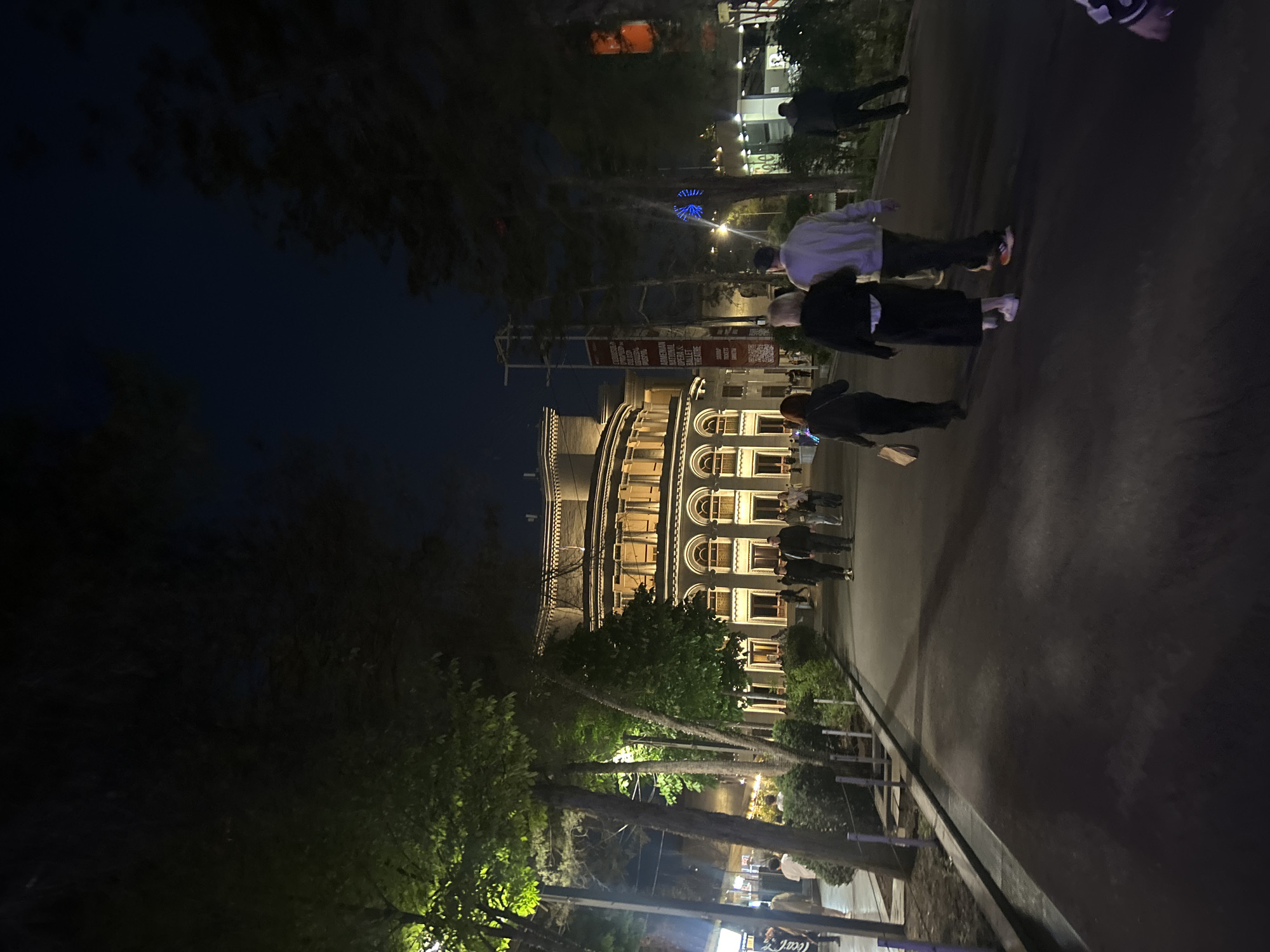 The image depicts an urban scene at night, taken from a slightly elevated perspective overlooking a paved street or pedestrian area. The dominant subject is a grand, multi-storied building in the middle ground, likely a theatre or opera house, illuminated with warm lights that highlight its classical architecture, arched windows, and decorative elements.
Several people are visible, walking casually along the paved area. In the foreground, figures are seen from behind, including individuals wearing a light-colored hoodie and dark pants, and another in a black top and white shoes. Further back, closer to the illuminated building, more pedestrians are present.
To the left of the paved area, trees and foliage line the street, with some receiving light from streetlights or nearby sources, creating a luminous green effect against the dark sky. In the deeper background, behind the trees and structures, a distinctive blue glowing object, possibly a decorative light installation or a Ferris wheel, is visible.
Banners are hanging vertically from poles near the illuminated building. One banner clearly displays "AMERICAN NATIONAL OPERA & BALLET THEATRE," along with "QUEEN" and "ROCK BALLET," indicating a performance. A QR code is visible on an adjacent banner. In the bottom right corner, a "Coca-Cola" logo can be faintly seen.
The time of day is clearly night, with a very dark sky and reliance on artificial lighting throughout the scene. The weather appears clear. The overall atmosphere is calm, suggesting an evening out or a leisurely stroll in a public square or boulevard, likely in Yerevan, Armenia.