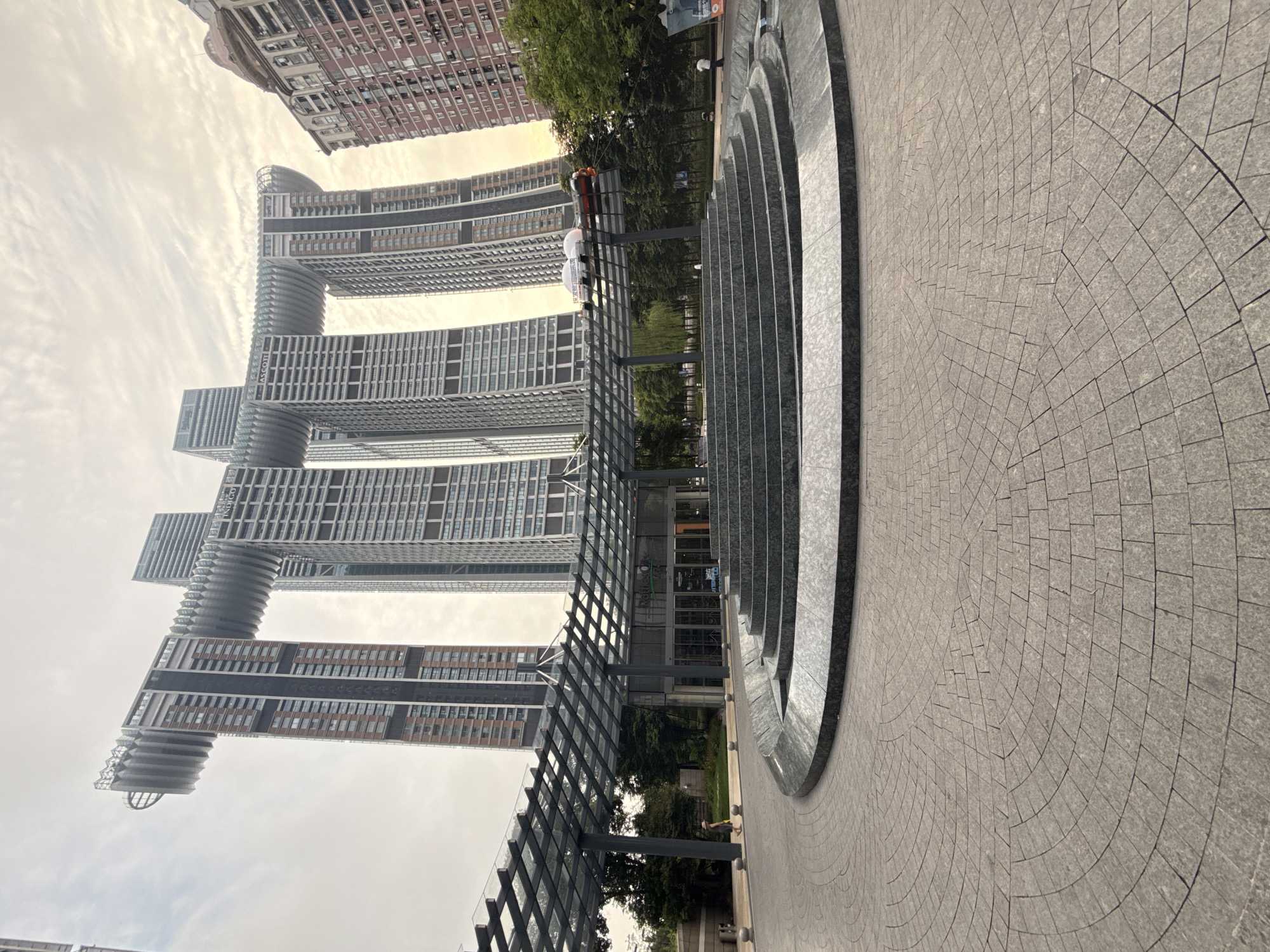 The photograph captures the exterior of the Raffles City Chongqing, a distinctive architectural complex located in the Yuzhong District of Chongqing, China. The dominant feature is the skyscraper complex, characterized by its unique "horizontal skyscraper" design, a connecting glass-and-steel structure that spans between multiple towers. These towers, clad in brick and glass, rise high into a hazy sky.
In the foreground, a circular plaza paved with interlocking grey stone tiles leads towards a tiered, circular water feature. This water feature appears to be a dry fountain or decorative basin. Above and behind the fountain, a transparent, angled roof structure supported by dark metal beams covers an entrance area to the buildings. The entrance has glass doors and signage, with the text "Raffles City" visible.
Surrounding the buildings and plaza are green trees and a few scattered individuals. One person, dressed in a light-colored outfit and hat, is walking on the paved pathway to the left of the fountain. Another person in a white shirt is standing near a tree to the right. The overall atmosphere is overcast, suggesting a diffused light condition. The architectural style is modern, with the large, interconnected buildings forming a striking urban landmark.