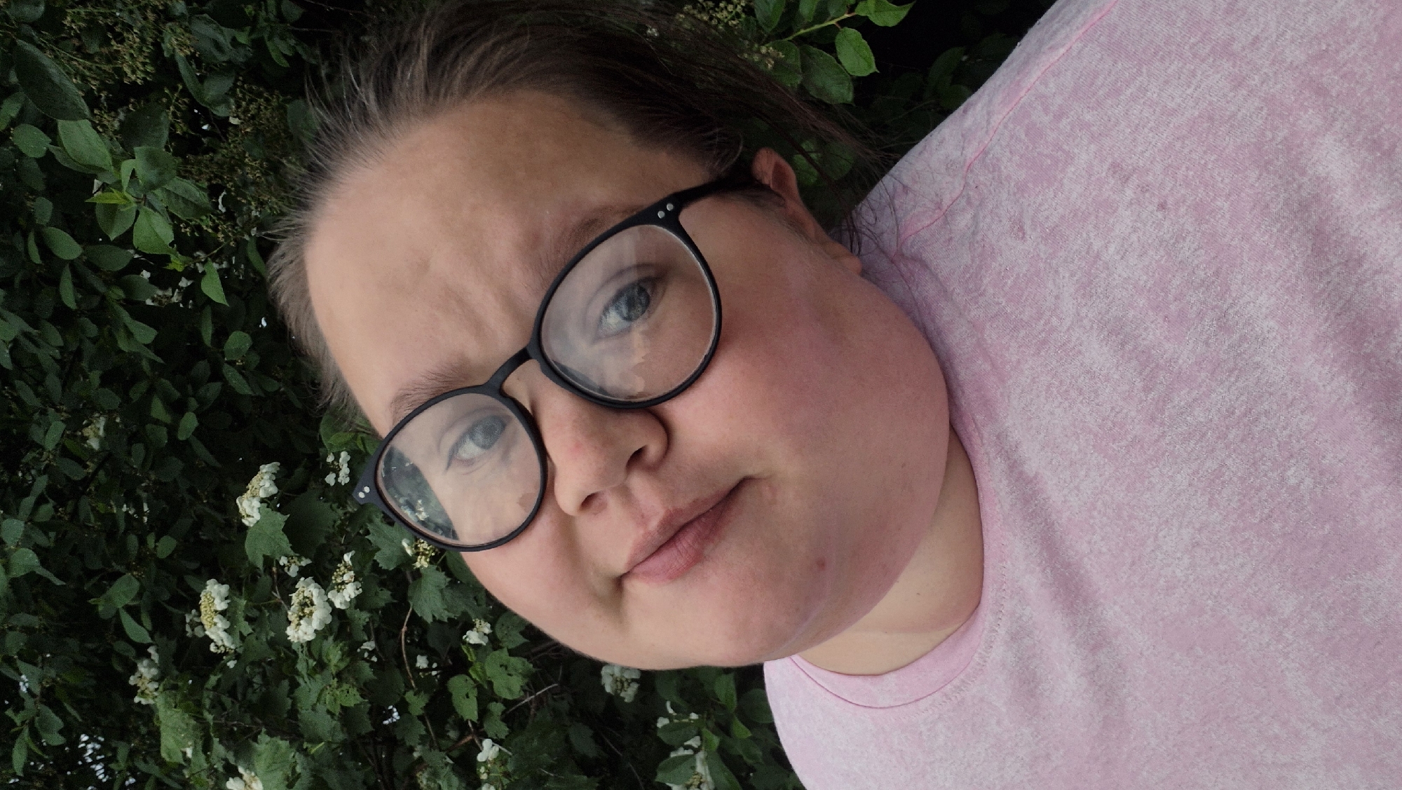 A selfie shows a fair-skinned woman with blue eyes wearing black-rimmed glasses and a light pink t-shirt. The woman’s face fills most of the frame, and she appears to be outdoors, with green foliage visible in the background, specifically leaves and small white flowers, possibly viburnum blossoms. The location is Chaikovsky, Russia.