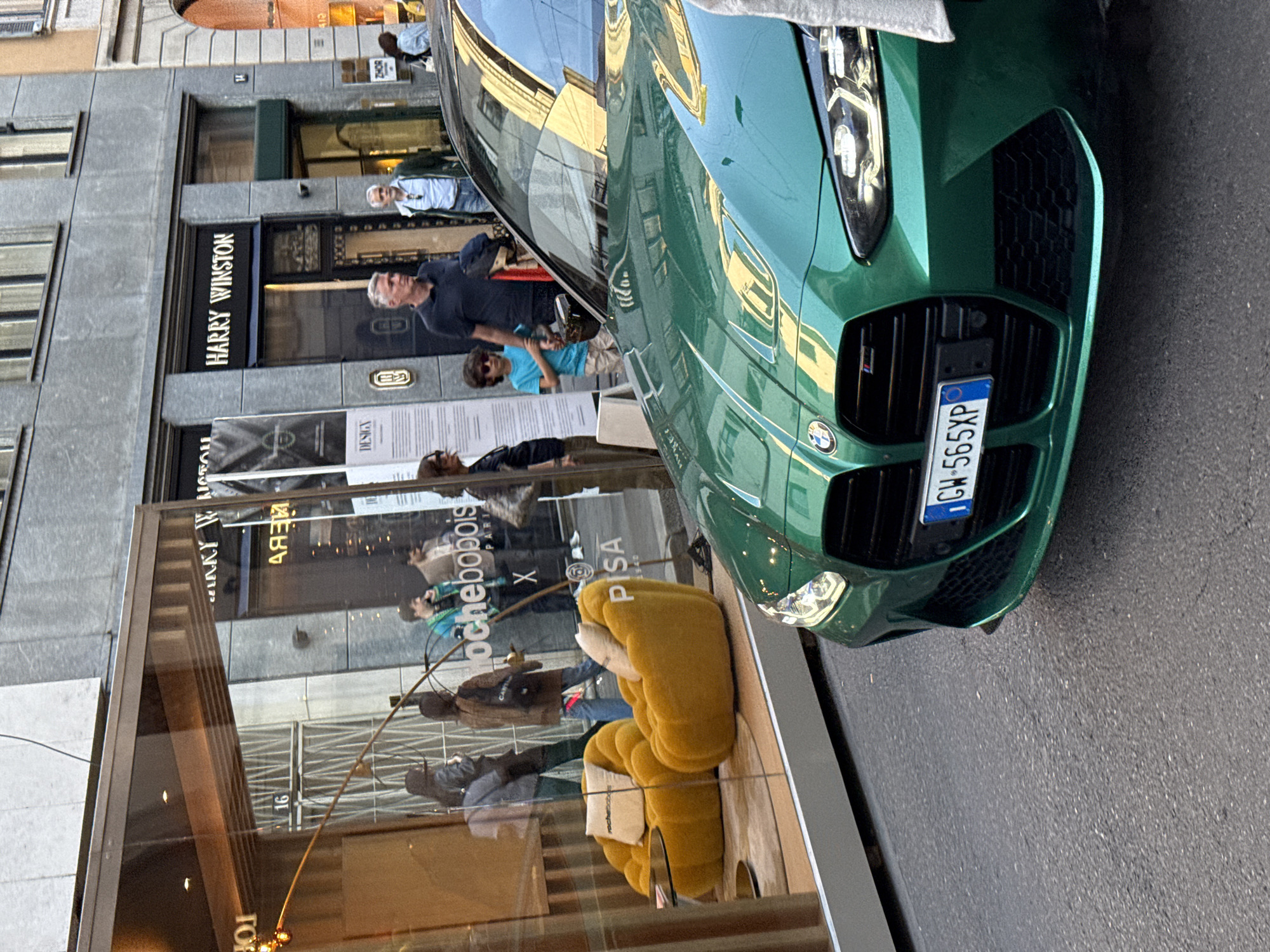 The image depicts a green BMW automobile parked on a city street, likely in Milan, Italy, based on the "CW" prefix on the license plate. The car is positioned in front of a storefront window, which reflects people and the surrounding architecture. Through the window, Roche Bobois furniture is visible, specifically a yellow sofa. The storefront is located next to Harry Winston, a luxury jeweler, according to signage. Several people are present: some reflected in the storefront window and others walking on the street. The photograph captures an urban scene that juxtaposes luxury goods with the everyday flow of city life.