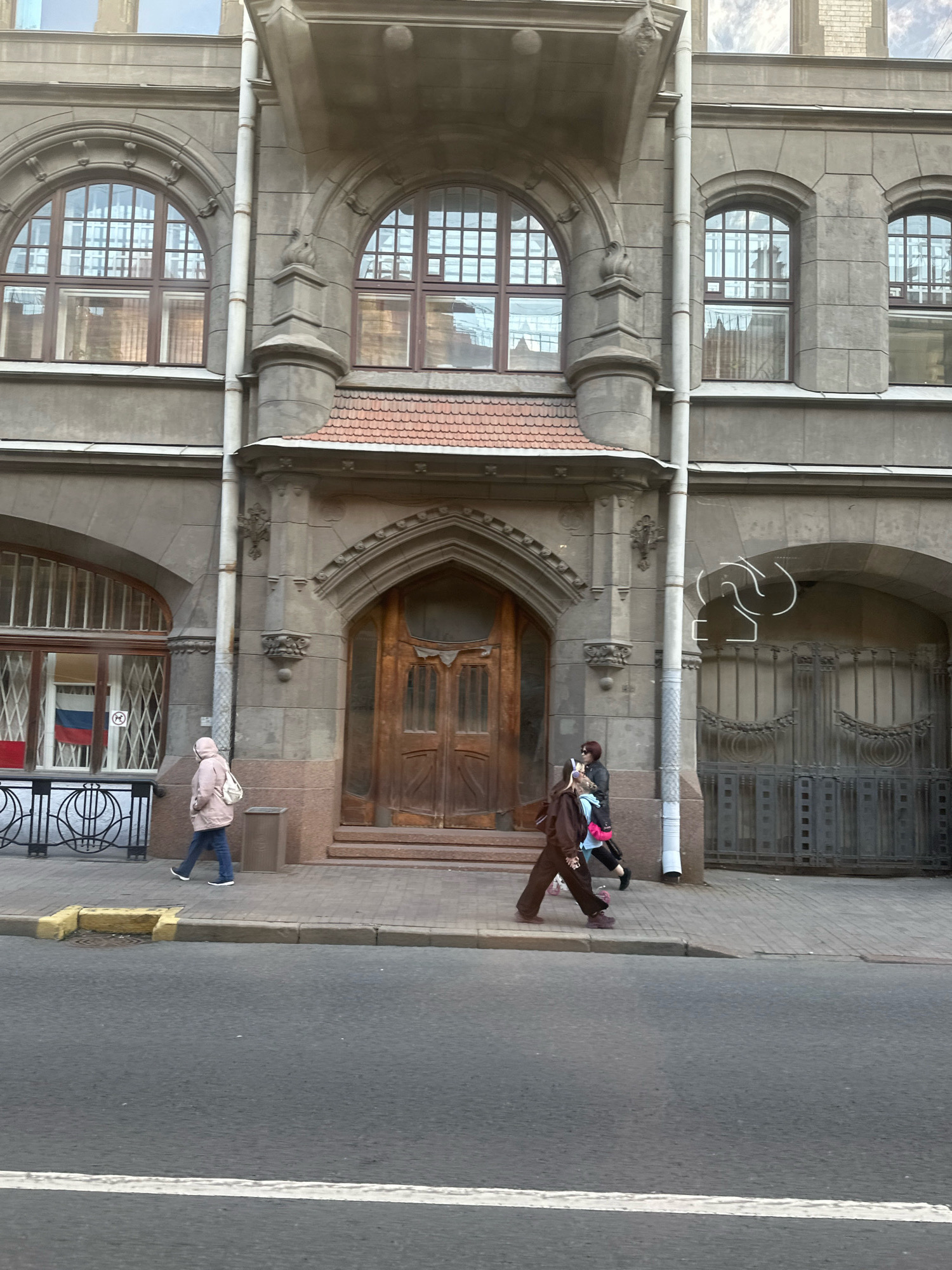 The image shows a street view of an old stone building in Saint Petersburg, Russia. The building has arched windows with ornate detailing and a large, wooden double door. A sidewalk runs in front of the building, and a road is in the foreground. Two women are walking on the sidewalk. The woman on the left is wearing a pink hooded jacket and blue jeans, carrying a white backpack. The woman on the right is wearing a brown hooded outfit and black pants, carrying a bag over her shoulder. She appears to be talking to another woman in a blue jacket and black pants who is partially obscured. In the window to the left, a Russian flag is visible. A large, decorative metal gate is visible on the right side of the building, and a white abstract line drawing is superimposed on the image above the gate.