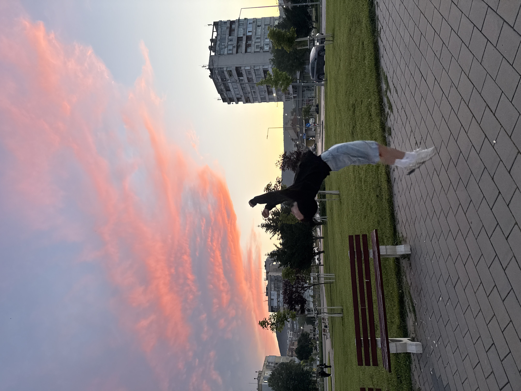 The image depicts an outdoor urban scene in Shumen, Bulgaria, during either sunset or sunrise. The sky is a prominent feature, showcasing a gradient of soft yellow and light blue near the horizon, transitioning into vibrant pink and fiery orange clouds across the upper portion of the frame, indicating the early or late hours of the day.
At the center of the composition, a person is captured mid-air, performing an acrobatic maneuver such as a backflip or parkour stunt. They are in a curved, inverted pose, wearing a dark long-sleeved top, light-colored shorts or pants, and white shoes.
In the foreground, there is a paved pathway made of grey rectangular bricks, running parallel to a lush strip of green grass. A dark red slatted park bench with light grey concrete supports is positioned on the grass next to the pathway.
The background reveals an urban environment with several multi-story apartment buildings, characteristic of Eastern European architecture, some appearing weathered with visible balconies. Trees are scattered throughout the park area and among the buildings. A streetlight and a parked car are also discernible. In the distance, a faint silhouette of mountains or hills can be seen against the horizon. A road sign in the background displays text in Cyrillic, with "БОРСА" (BORSA) partially visible, confirming the Bulgarian setting.
The image captures a dynamic human activity set against the striking beauty of a colorful sky and an urban landscape.