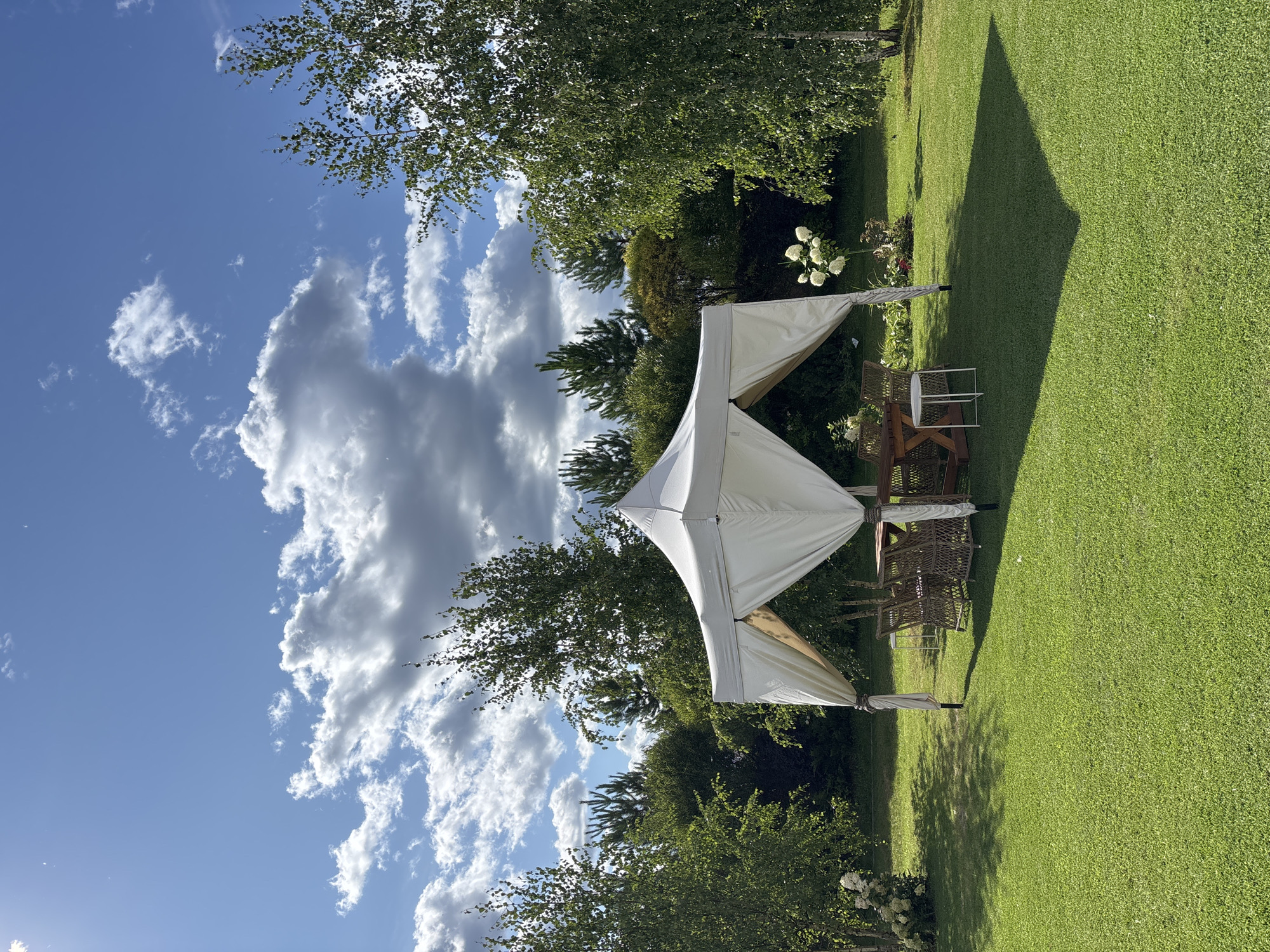 The image depicts a sunlit outdoor scene in Pokrovskoe, Russia. A white canopy tent is set up on a lush green lawn, casting a shadow. Beneath the tent, a wooden table is surrounded by wicker chairs. To the left of the tent, white flowers are in bloom, and to the right, a birch tree stands tall. In the background, a dense line of various trees creates a natural backdrop against a bright blue sky filled with white, fluffy clouds. The overall impression is one of a peaceful, natural setting, likely a garden or park, during a clear and sunny day.