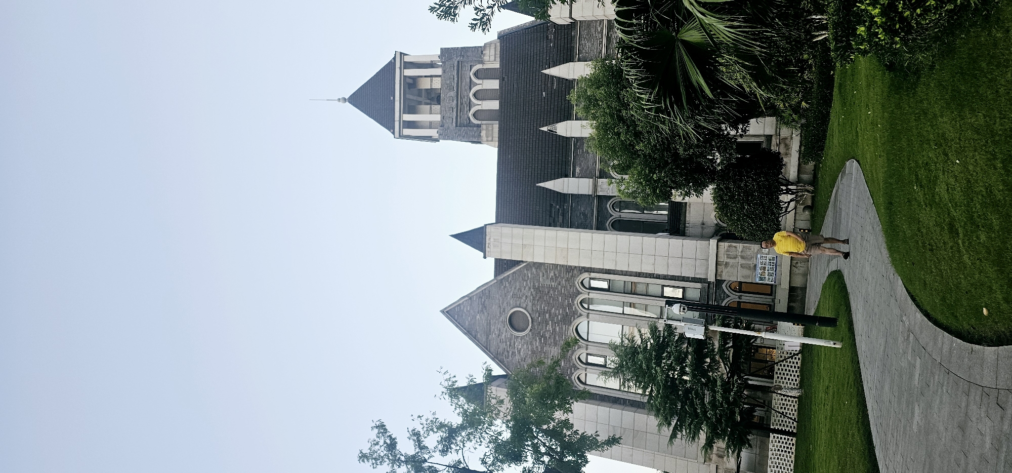 A man in a yellow shirt and khaki shorts walks along a curved, paved path in front of a large, gothic-style stone building. The building has a tall, pointed tower with arched windows and a dark, pitched roof. Lush green grass and trees surround the path and the building. The sky is overcast and pale blue. The scene is located in the Chongqing High-tech Zone, China.