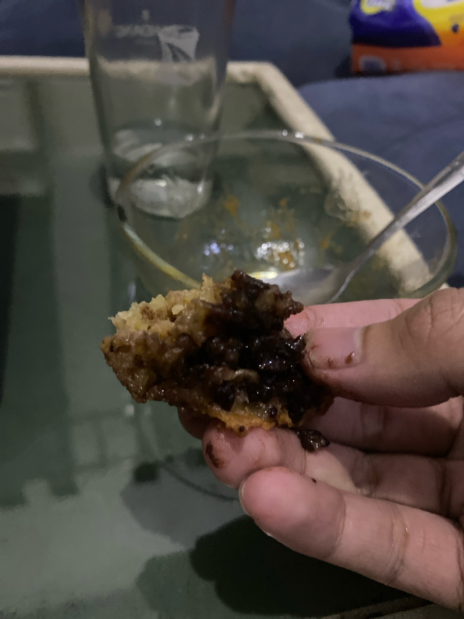 A close-up, handheld shot shows a person's fingers holding a piece of food that appears to be fried and filled with chocolate. The food is golden brown and appears somewhat crispy on the outside, with dark, melted chocolate visible within. The person's fingers are slightly greasy, suggesting they have been eating the food. In the blurred background, a glass of water and a bowl are visible, as well as what looks like a couch and a colorful package of snacks. The scene is indoors, likely in a residential setting in Ngalian, Indonesia, possibly during a casual meal or snack time. The lighting is dim, casting shadows and creating a cozy atmosphere.