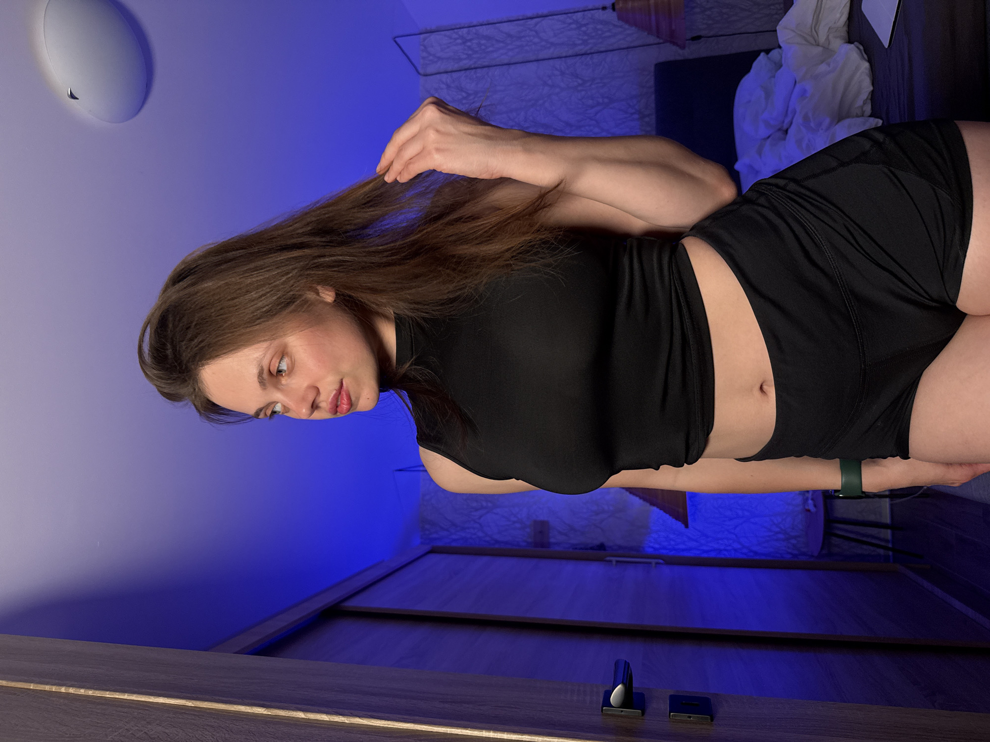 The image depicts a young woman standing in a dimly lit room, with a strong blue light casting shadows. She is wearing a black sleeveless crop top and black shorts, with a green smartwatch on her left wrist. Her hair is brown and long, and she is looking to her left, with her right hand touching her hair. The room appears to be a bedroom, with a bed and a wardrobe visible in the background. The overall atmosphere is intimate and slightly moody due to the lighting. There is no information in the image that suggests the location is the city of Vrchlabí, Czechia.