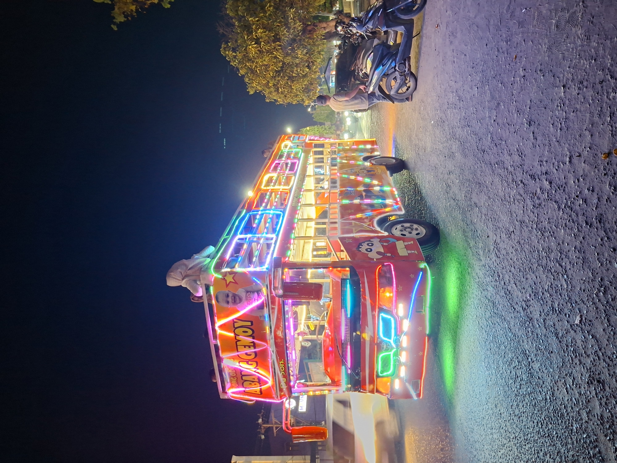 The image captures a brightly illuminated "ODONG ODONG" ride on a street in Lhokseumawe, Indonesia, at night. This form of ride usually carries children and is usually decorated with bright lights and characters. The vehicle is a makeshift, open-air bus adorned with neon lights of multiple colors. The ride's name is “ABAK CEMMOY SON.”
At the edge of the frame, on the sidewalk near the ODONG ODONG ride, some motorcycles are parked. Several other vehicles are in the background along the street. In the background, one can see a street lamp and some trees and buildings.