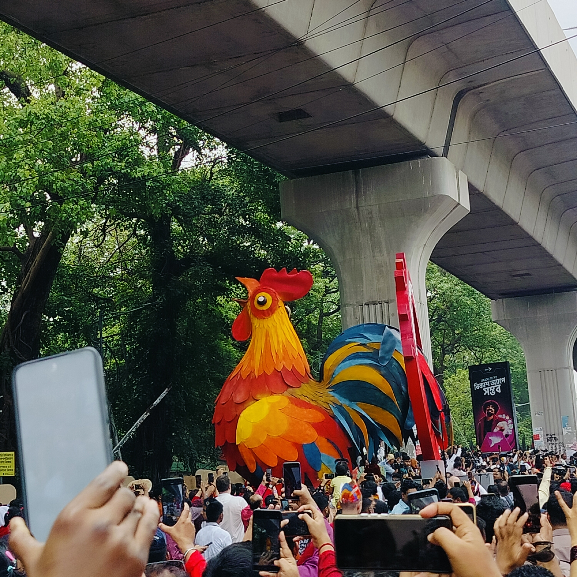 A large, colorful rooster effigy is the focal point of an outdoor scene in Dhaka, Bangladesh. The effigy has a red crest, yellow and orange body, and blue and yellow tail feathers. It is surrounded by a large crowd of people, many of whom are holding up their cell phones to take pictures. The effigy appears to be stationary. Above the crowd and effigy is a large, concrete overpass. Lush green trees are visible on the left side of the frame. A banner with a portrait and writing in Bengali can be seen behind the effigy. The scene suggests a festival, parade, or public celebration.