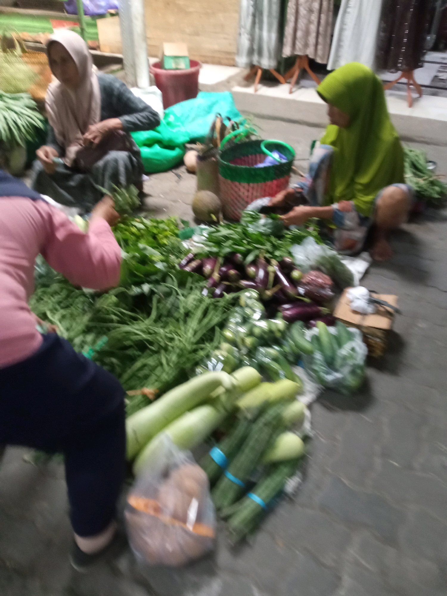 The image depicts an outdoor market scene, likely in Balun, Indonesia, characterized by a concrete ground and stalls with various fresh produce. Two women, dressed in hijabs, are present. One woman, wearing a light pink hijab and a dark gray top, is seated on the ground, sorting through a pile of green leafy vegetables. The other woman, wearing a bright green hijab, is also seated, handling vegetables. A variety of fresh vegetables, including what appear to be eggplants, gourds, cucumbers, and leafy greens, are spread out on the ground. Several woven baskets and plastic bags are visible, containing or holding the produce. In the background, a red bucket and some fabric displays suggest a marketplace setting. The overall lighting is dim, indicating it might be evening or an indoor market with artificial light. The focus is on the produce and the sellers actively engaged in their work.