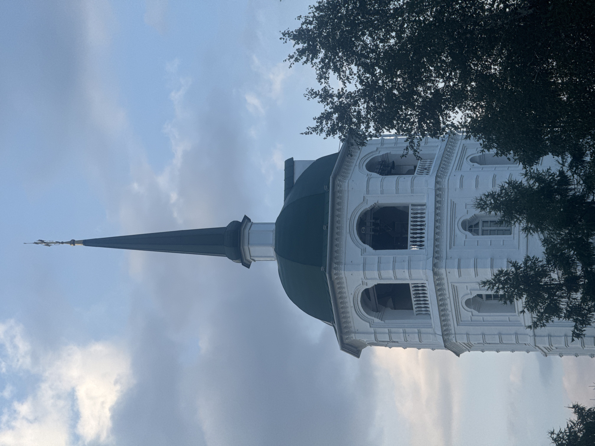This is a close-up, medium-shot, vertical, outdoor photograph of the white bell tower of the Epiphany Cathedral in Irkutsk, Russia. The bell tower has a green domed roof, an octagonal shape, and multiple levels with arched windows and balconies. The sky is partly cloudy, with a mix of blue and gray clouds. Dark green evergreen trees are visible in the foreground, partially obscuring the lower part of the bell tower. The top of the bell tower culminates in a dark green spire topped with a cross. The overall lighting suggests it is either early morning or late afternoon. There are no people or animals visible in the image.