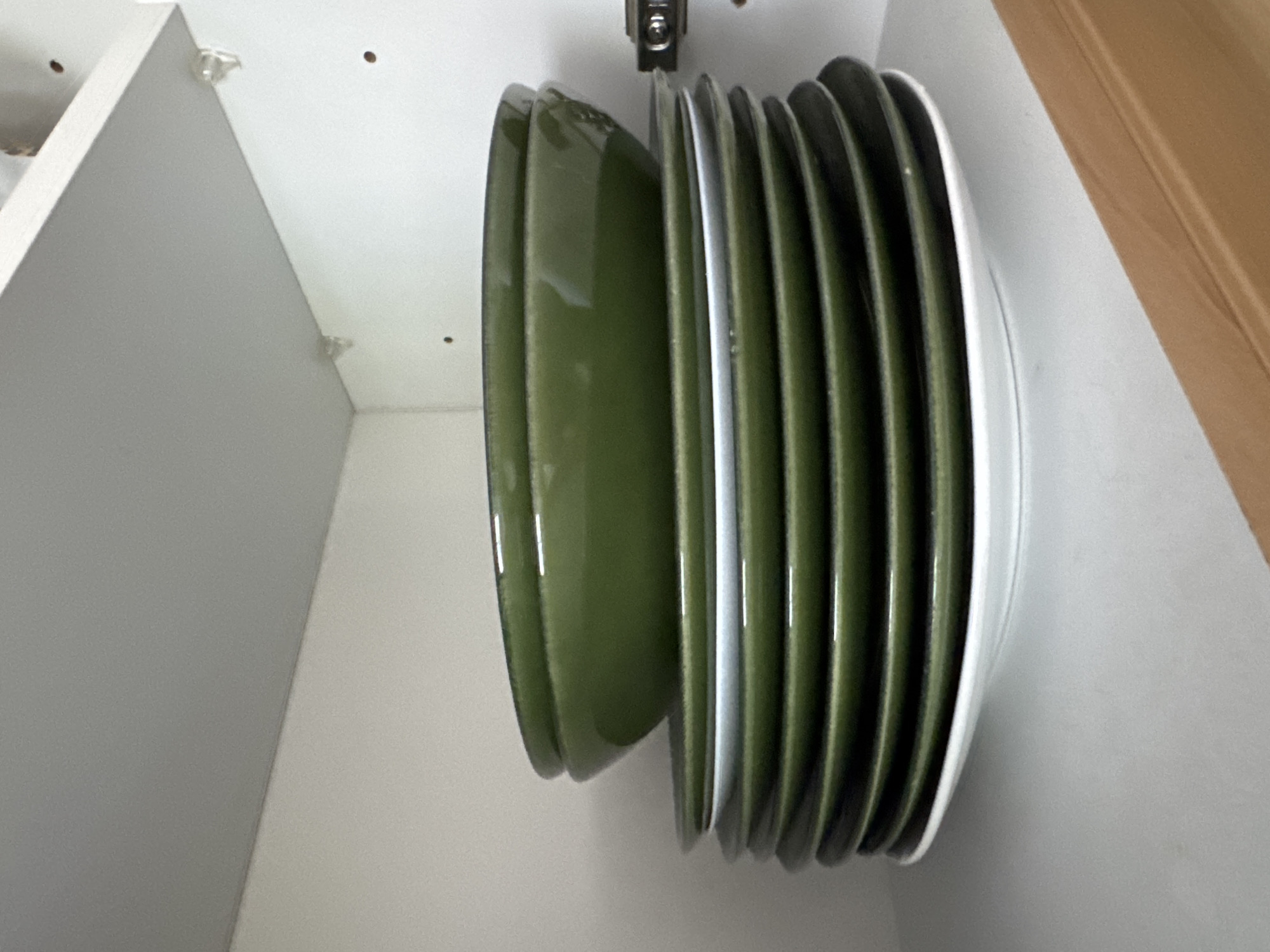 A stack of green bowls and white plates is neatly arranged inside a white cabinet in a home in Höhenkirchen-Siegertsbrunn, Germany. The cabinet is open, revealing its interior. The bowls are stacked on top of the plates, with two large green bowls at the top, followed by a stack of several green plates, and then a single white plate at the bottom. The lighting suggests the photo was taken indoors during the day.