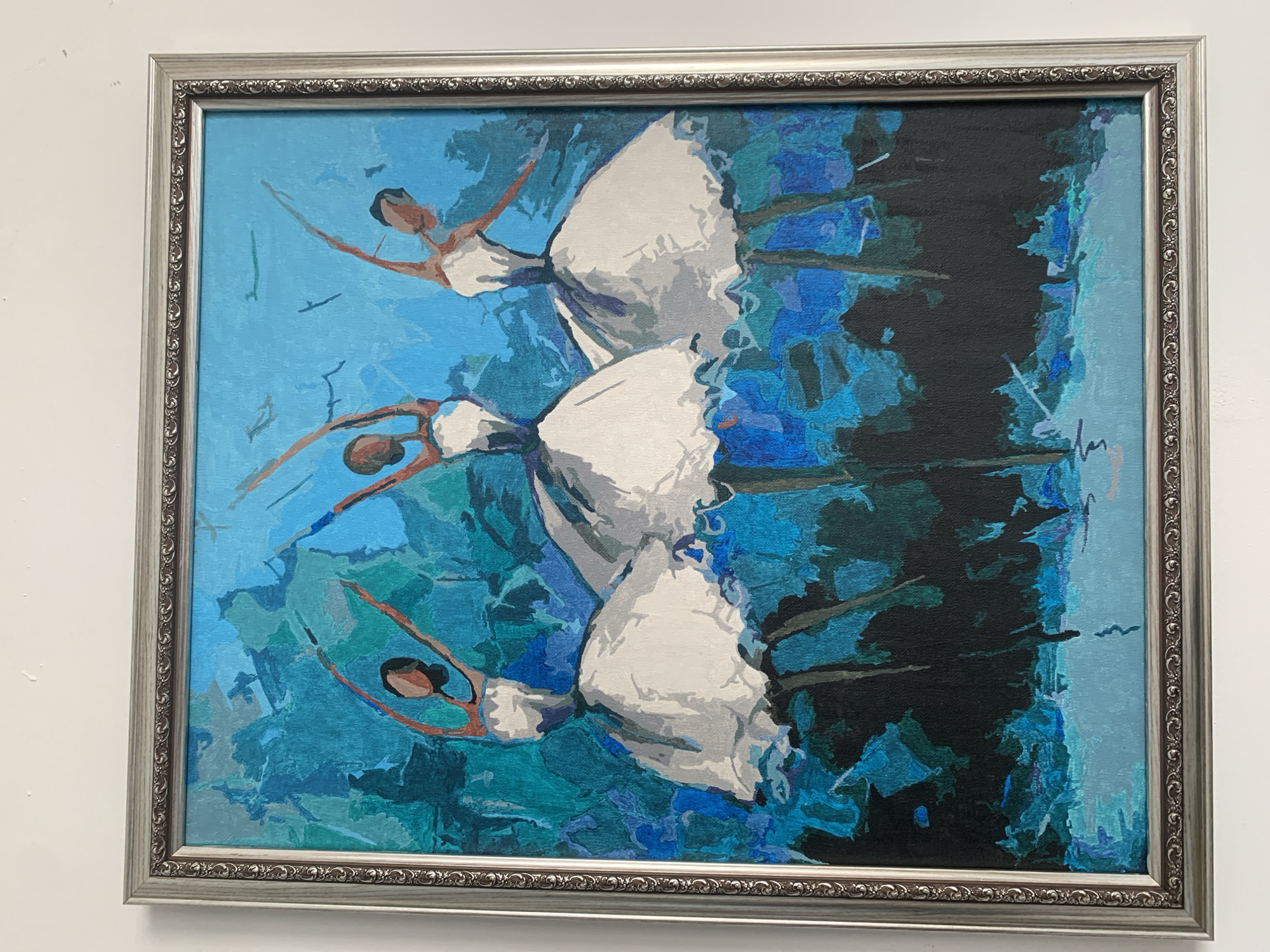 A framed painting hangs on a white wall in an unknown location in the city of Unknown. The painting depicts three ballerinas in white dresses against a blue and black background. The ballerinas are in varying poses, seemingly leaping or dancing. The artwork is executed in a style with visible brushstrokes and bold colors. The frame is silver with an ornate design.