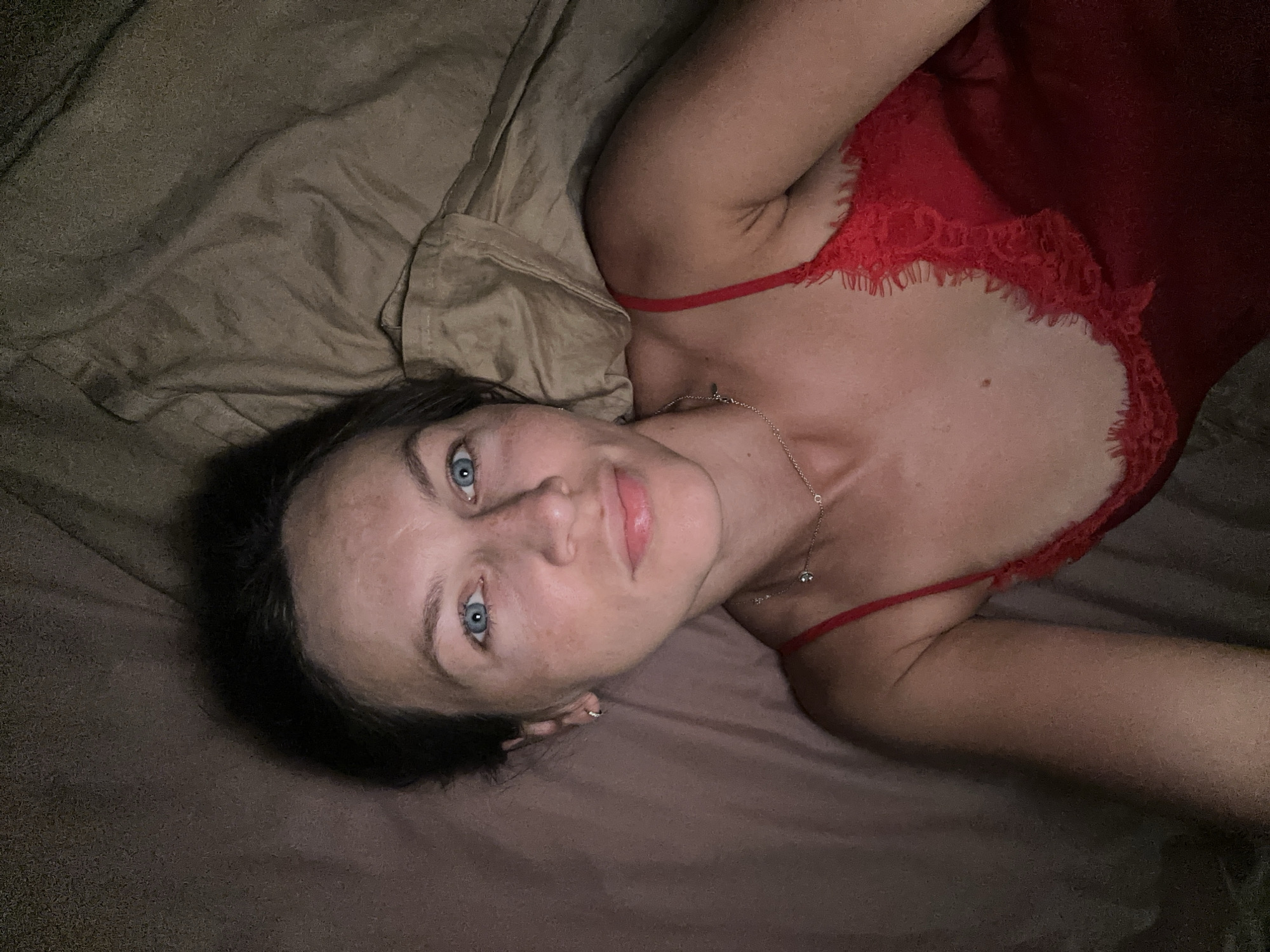 A woman with blue eyes and dark hair lies in bed, wearing a red lace-trimmed camisole. She is lying on her side, with her head tilted slightly upwards, facing the camera. The background consists of bedding in shades of brown and beige, with visible wrinkles and folds indicating it is a used bed. The lighting is dim, suggesting it is nighttime or a dimly lit room. There are no discernible urban or environmental features visible that would indicate a specific city location.
