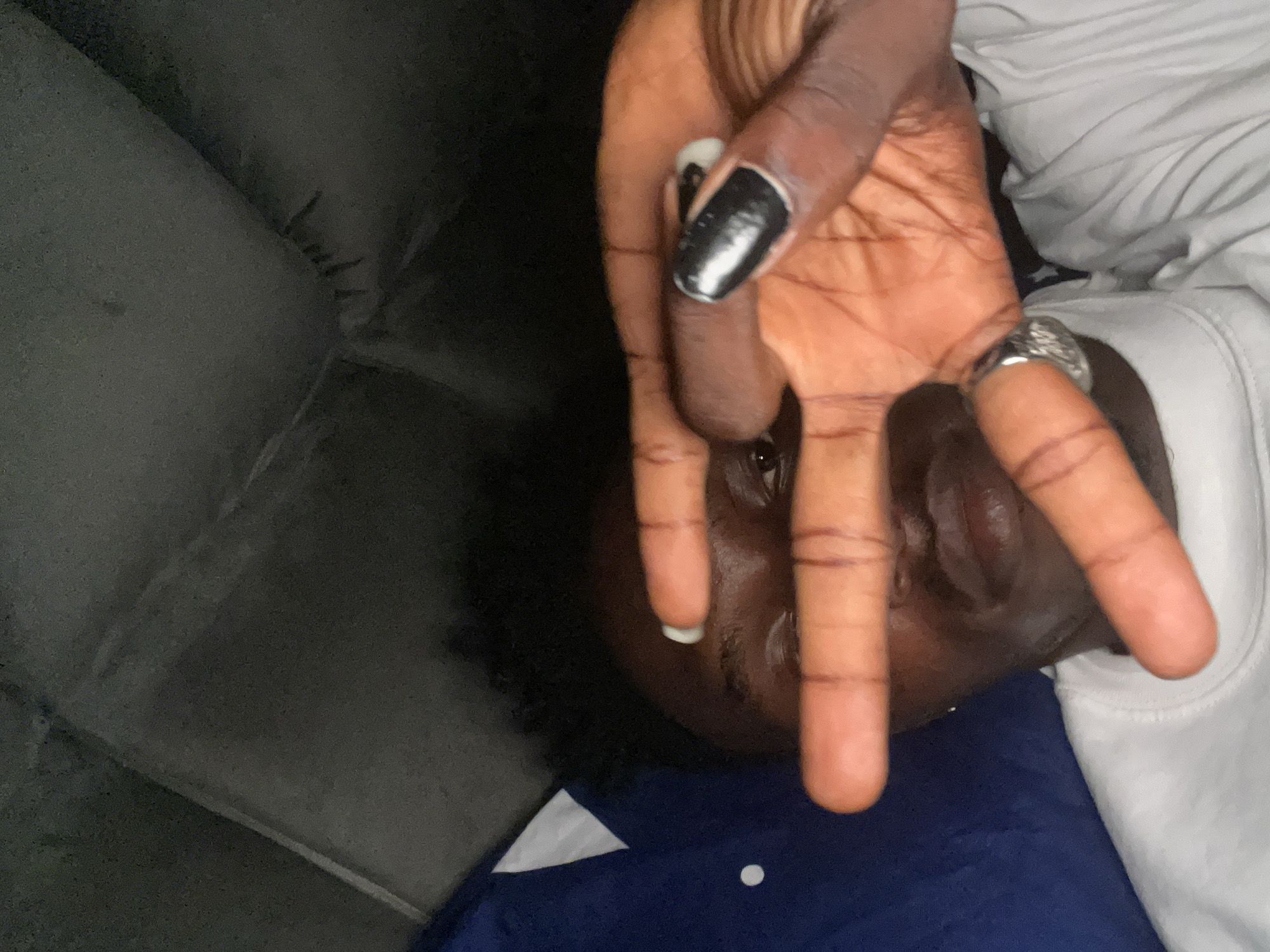 A person with brown skin lies on a gray surface, presumably a couch. The person is wearing a white shirt with a blue layer underneath. The person's right hand is raised, obscuring part of their face. The index finger is bent, the middle finger is raised and has a ring on it, and the thumb is bent and has black nail polish. The scene appears to be indoors. Lokoja, Nigeria cannot be determined from the image.