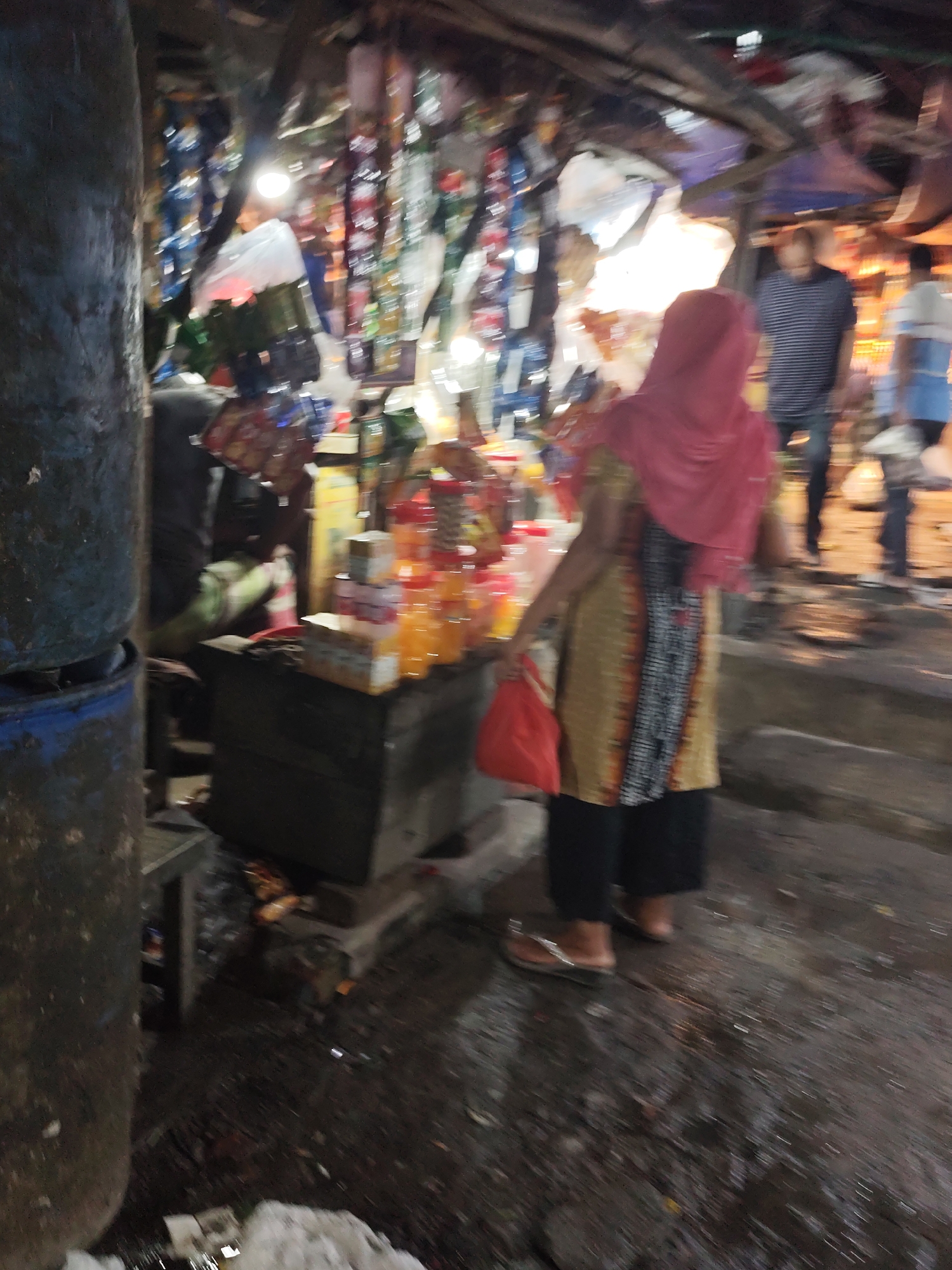 This image depicts a bustling street scene in what appears to be a market area in Dhaka, Bangladesh. A woman wearing a red headscarf and patterned clothing is standing near a vendor's stall. The stall displays stacks of food and beverages, including bottled drinks. The woman holds a red plastic bag, suggesting she is a customer who has just made a purchase. The foreground includes a large, possibly rusted, metal container. The ground is wet and littered with debris. In the background, two men can be seen walking away from the stall. The environment is somewhat dimly lit and crowded, characteristic of street markets in Dhaka.