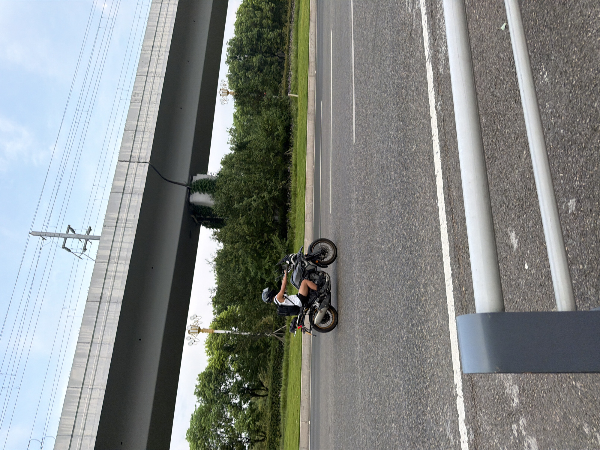 A person wearing a helmet and a white tank top rides a dark-colored motorcycle with a backpack on their back, traveling on a paved road. The motorcycle has a license plate that is not visible. The road has white lane markings. In the background, a large concrete overpass runs horizontally across the frame. Below the overpass, a line of green trees and grass is visible, and a lamppost with multiple lights stands near the trees. Overhead power lines are strung across the sky, supported by utility poles. The scene appears to be in a suburban or urban area, possibly on the outskirts of the city of Jiangjin, China, given the presence of overpasses and extensive infrastructure. The sky is partly cloudy. In the foreground, a metal guardrail runs horizontally.