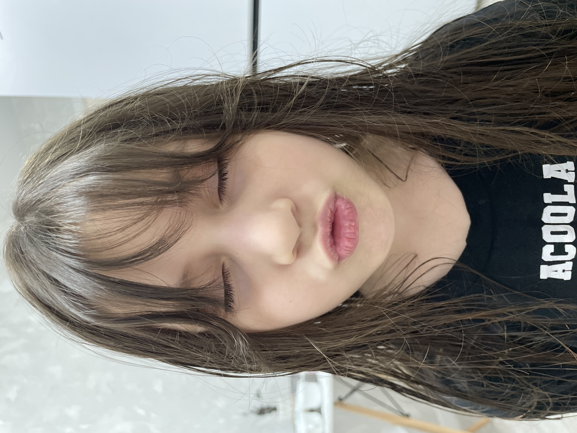 A young girl with brown hair and bangs, wearing a black t-shirt with white lettering, is lying down with her eyes closed. The scene appears to be indoors, possibly in a home setting. The location is not possible to ascertain based on visual details alone. There is no information about actions or interactions in the image.