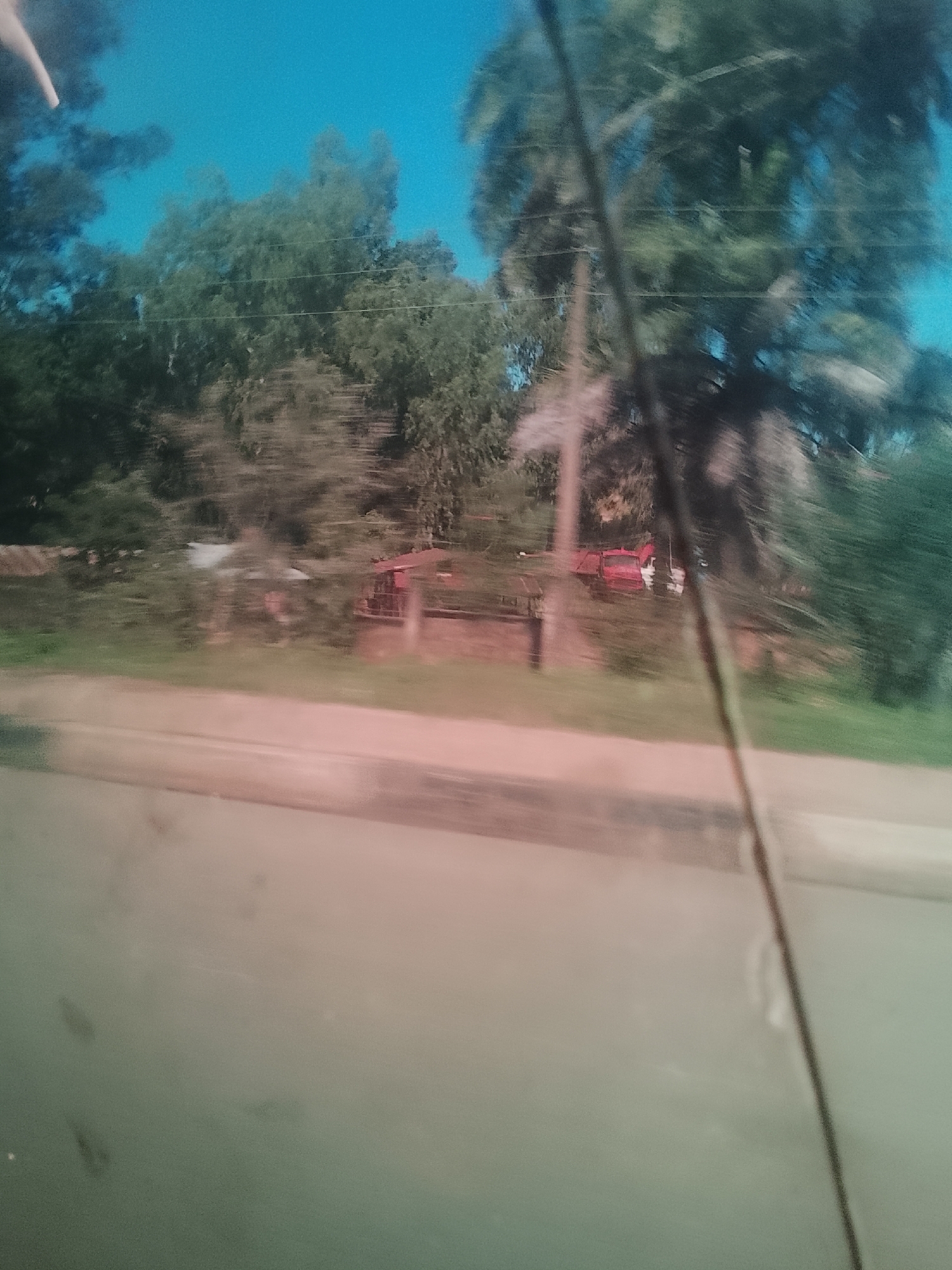 The image shows a blurry, outdoor scene with lush green trees and a bright blue sky. In the middle ground, there appears to be a red truck or vehicle, partially obscured by foliage. The foreground is a blurred expanse of what might be a road or a body of water, with a light pinkish hue. The overall impression is of a snapshot taken from a moving vehicle, capturing a fleeting glimpse of a rural or semi-rural environment. The location provided, Gura Top, Nigeria, suggests a specific geographical context, likely a town or region within Nigeria.