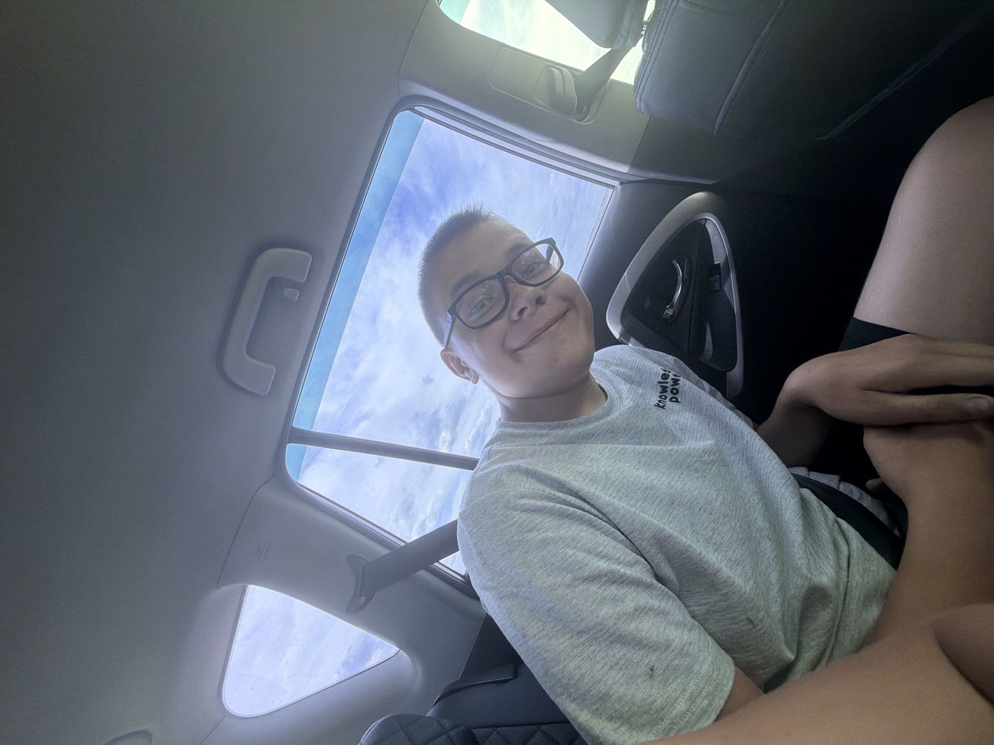 A young person with short brown hair and glasses, wearing a grey t-shirt, is seated inside a car. The t-shirt has black text on the front that reads "knowledge power". The person is looking towards the camera with a slight smile and is positioned in the back seat, with the car's interior visible, including a seatbelt, car door handle, and the black leather headrest of the seat in front. The sky with clouds is visible through the car windows, suggesting an outdoor daytime setting. The location is not discernible from the image.